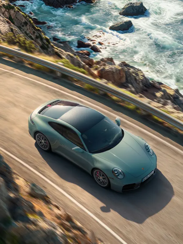 En lysegrøn Porsche 911 Carrera 4S ses oppefra, mens den kører på en kystvej ved havet.