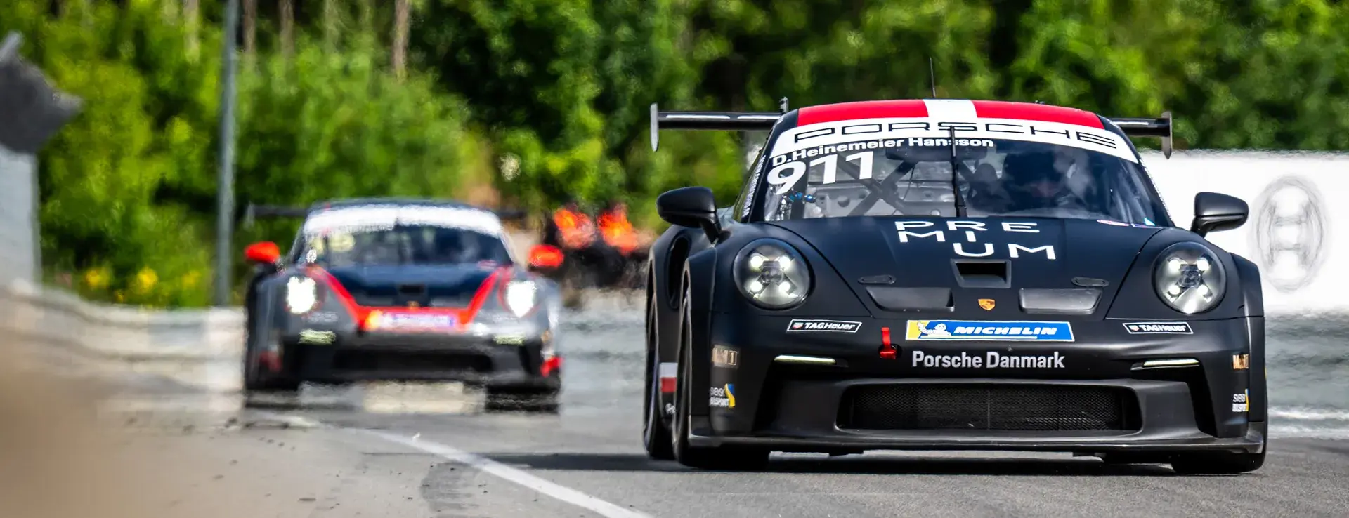 Sort Porsche-racer med nummer 911 i front på racerbane under Porsche Festival, med en anden bil tæt bagved.