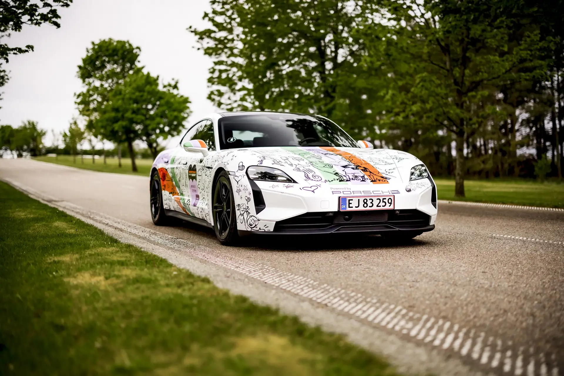 En hvid Porsche Taycan Turbo med farverig, håndtegnet udsmykning kører på en landevej omgivet af grønne træer.