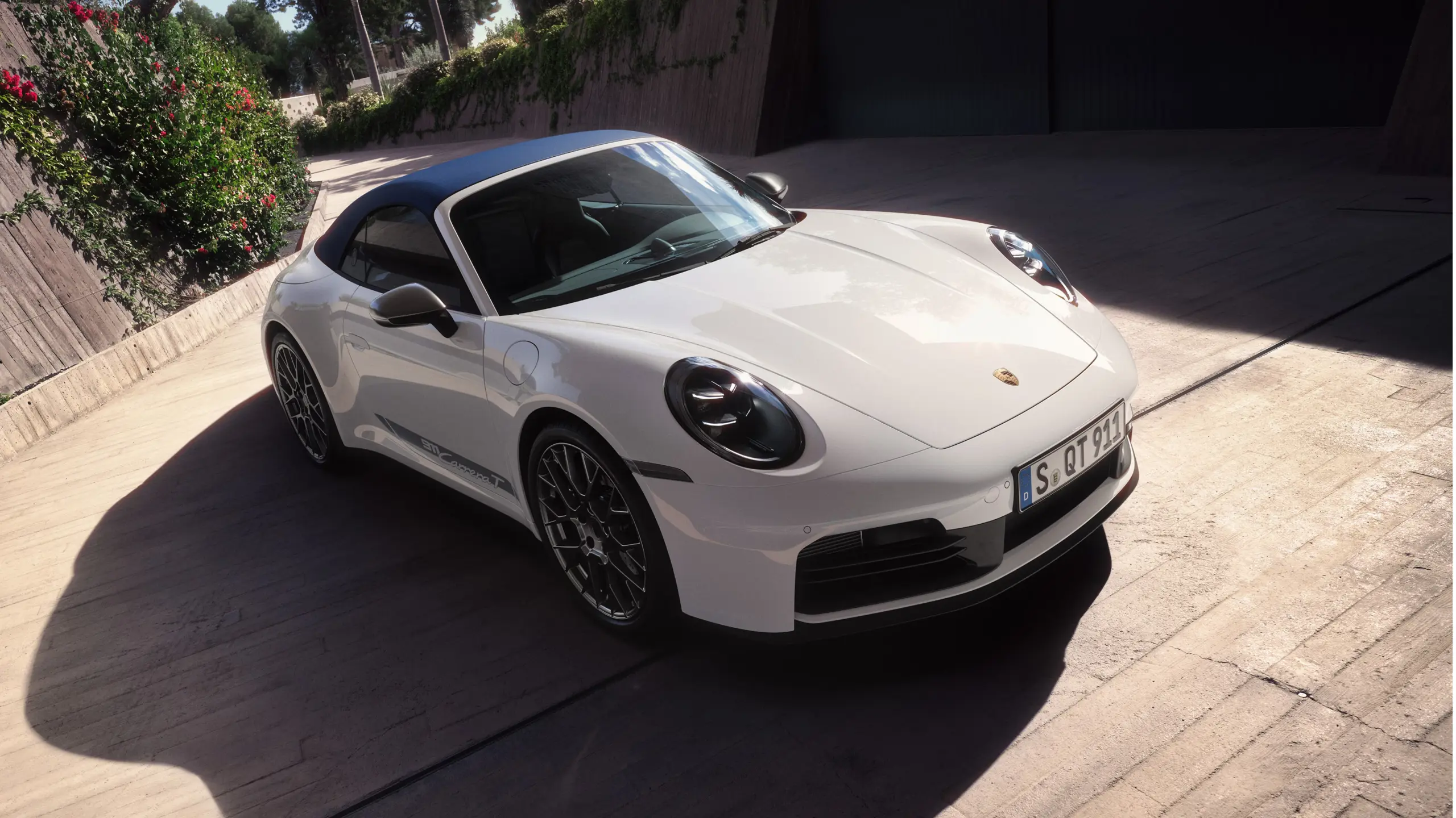 En hvid Porsche 911 Carrera T cabriolet holder parkeret udendørs foran en moderne bygning med grøn bevoksning på murene.