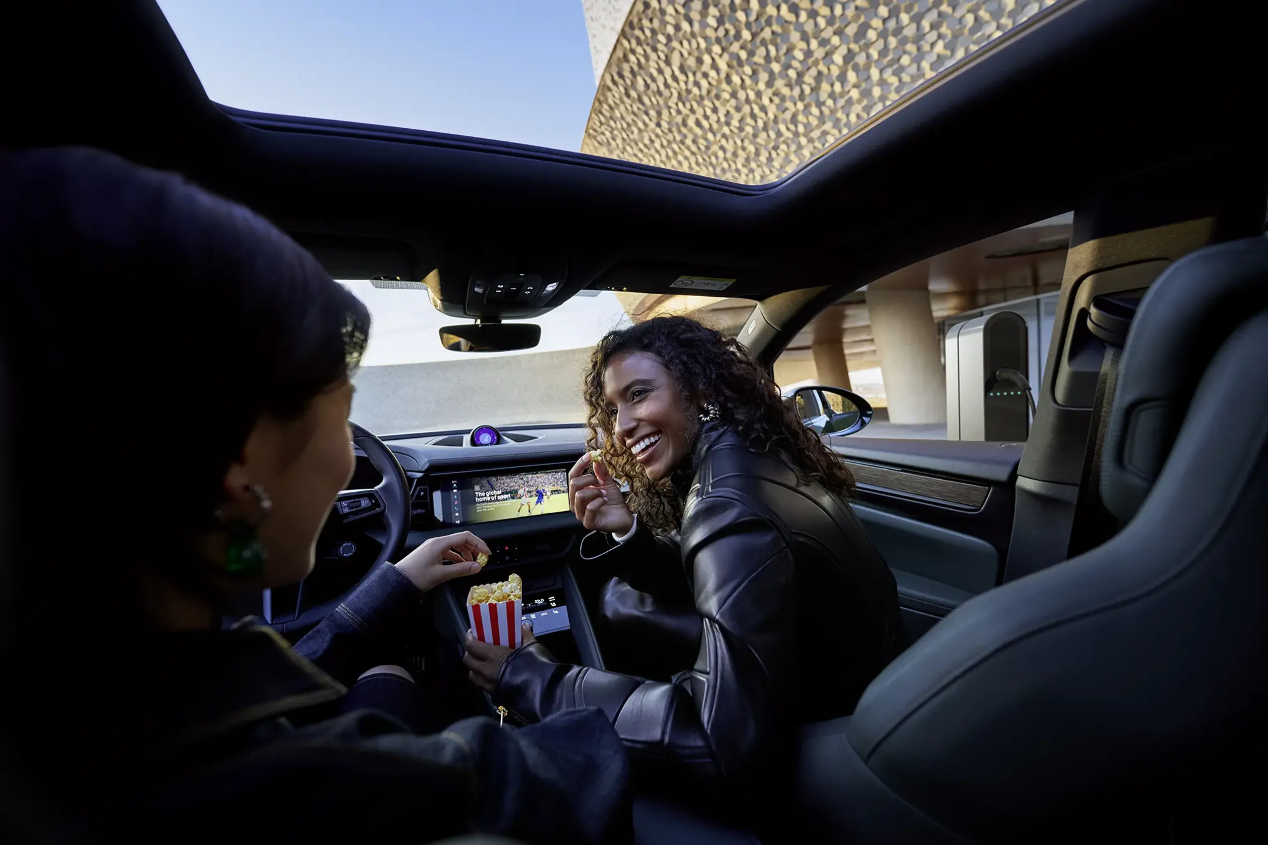 To kvinder sidder i en moderne Porsche, ser film på den centrale skærm og spiser popcorn, mens bilen lader ved en ladestander.