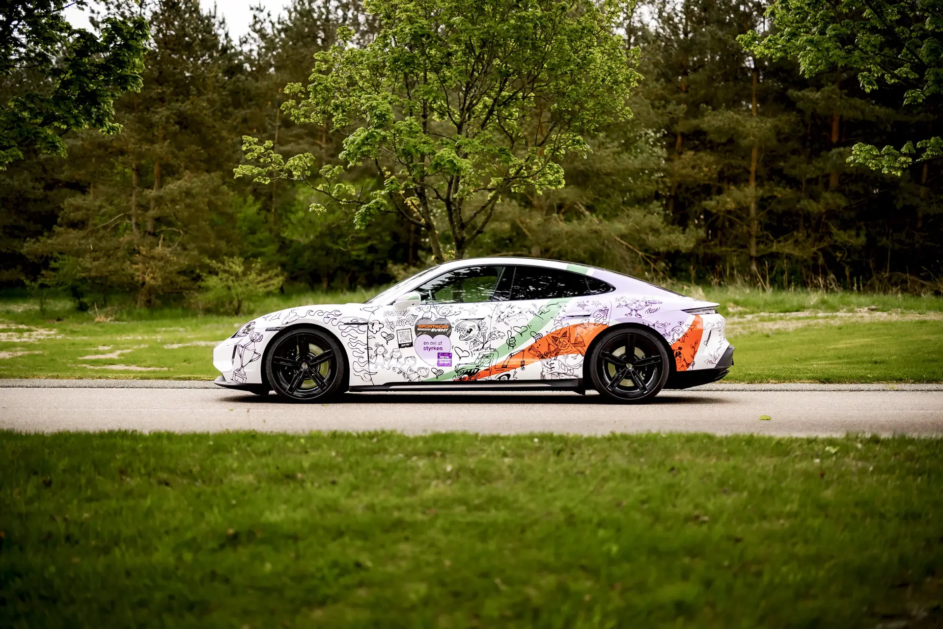 En hvid Porsche Taycan Turbo med farverig, grafitti-lignende folieparkering står på en vej omgivet af grønt græs og træer.