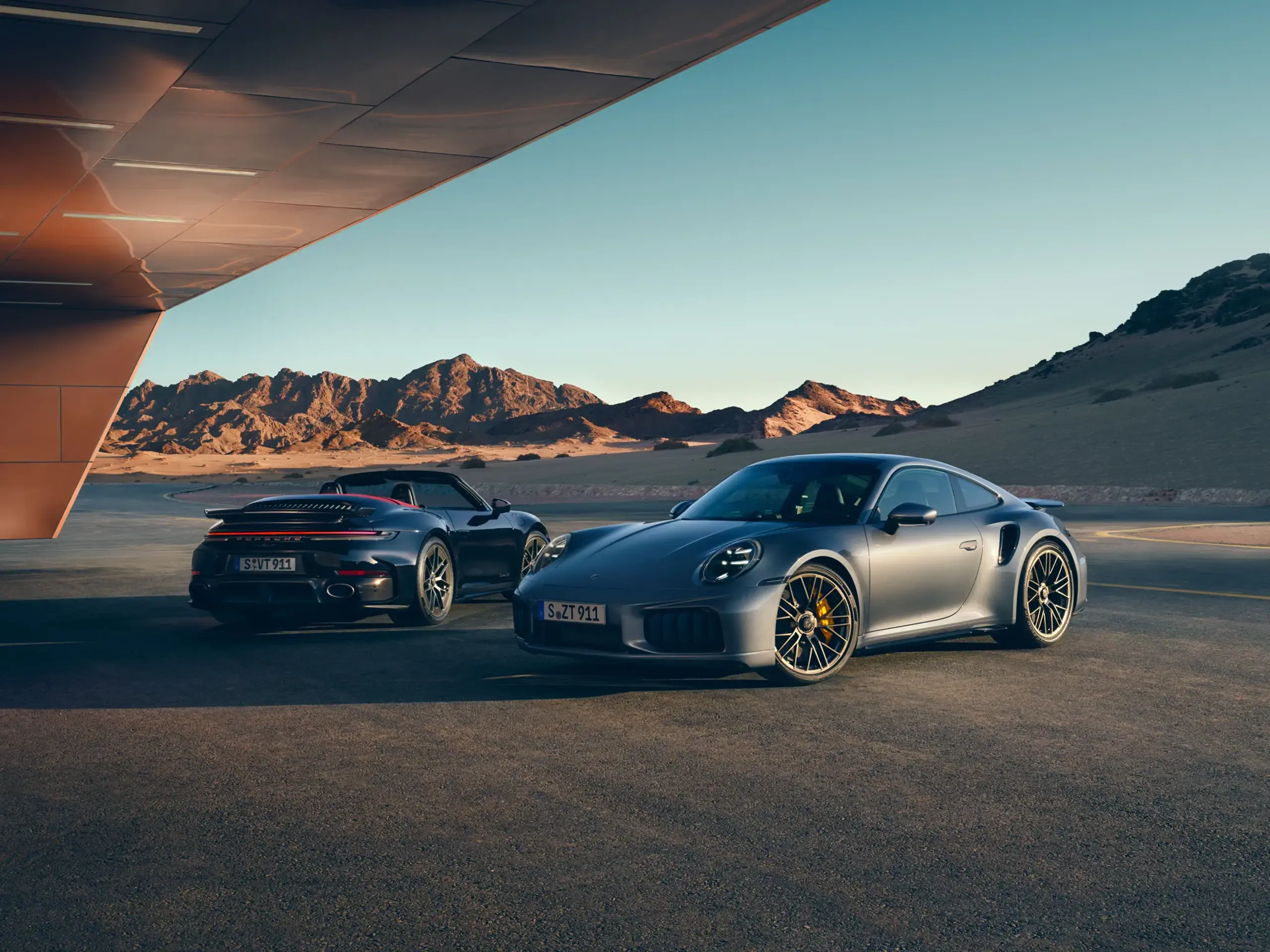 To Porsche 911 Turbo S sportsvogne holder parkeret i et ørkenlandskab ved solnedgang.