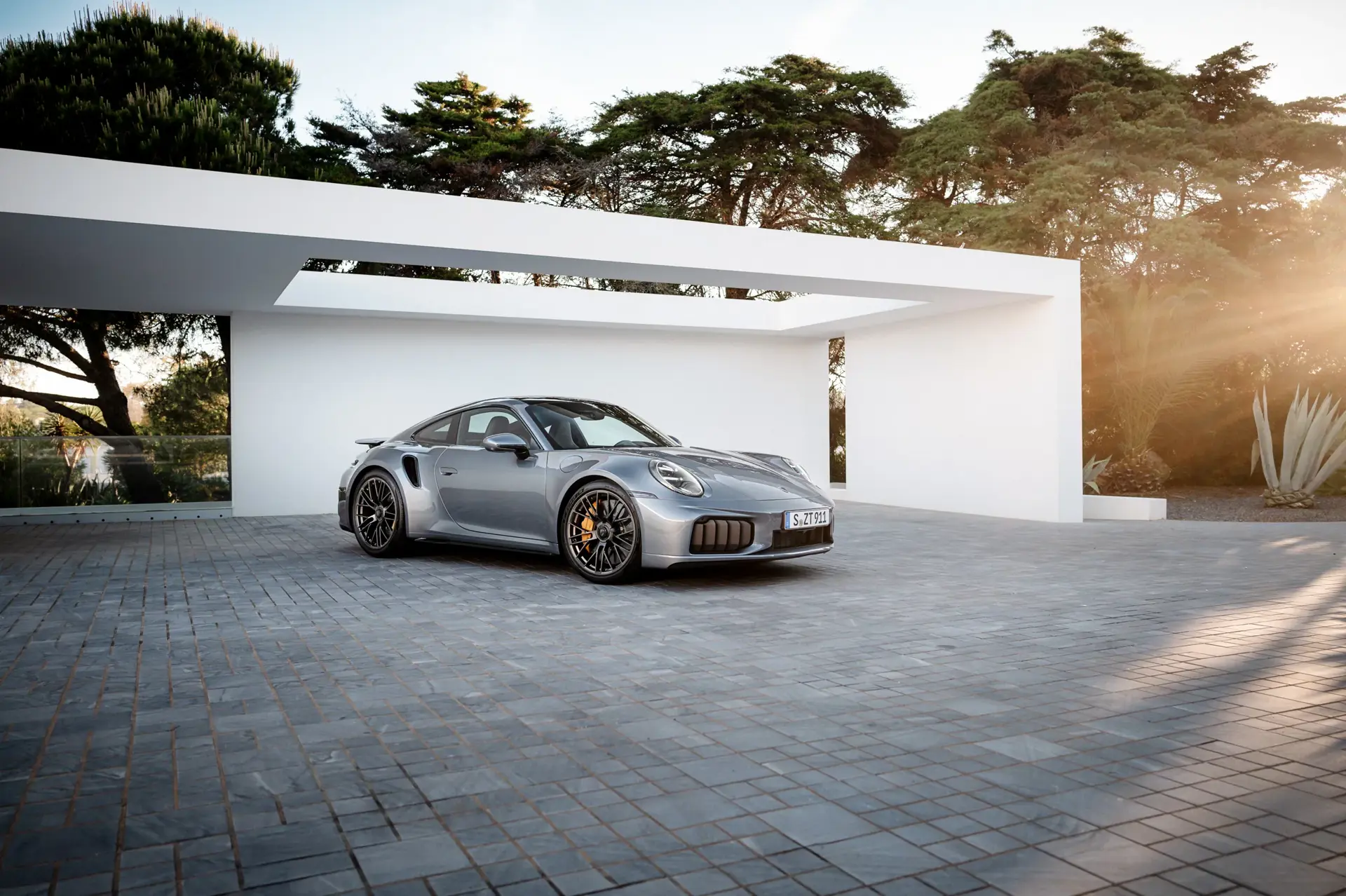 En sølvgrå Porsche 911 sportsvogn holder parkeret foran en moderne, hvid villa i solnedgangen.