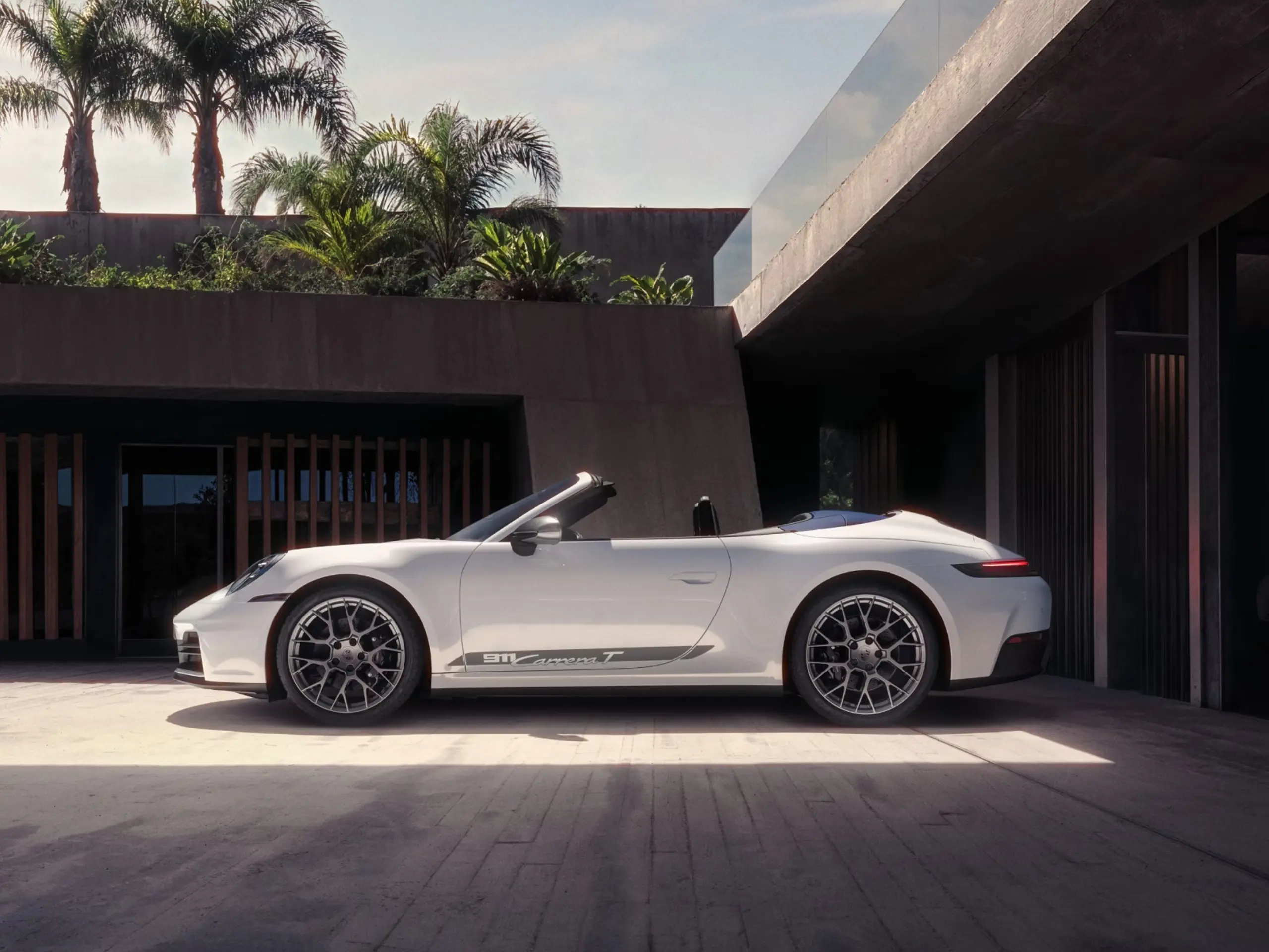 En hvid Porsche 911 Carrera T cabriolet holder parkeret foran en moderne betonvilla med palmer i baggrunden.