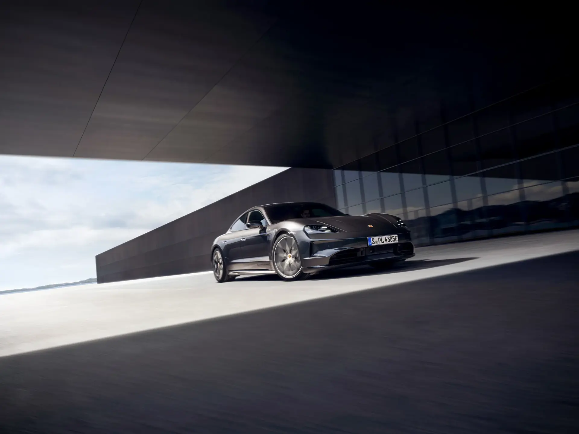 En mørkegrå Porsche Taycan ses i fart foran en moderne glas- og betonbygning.