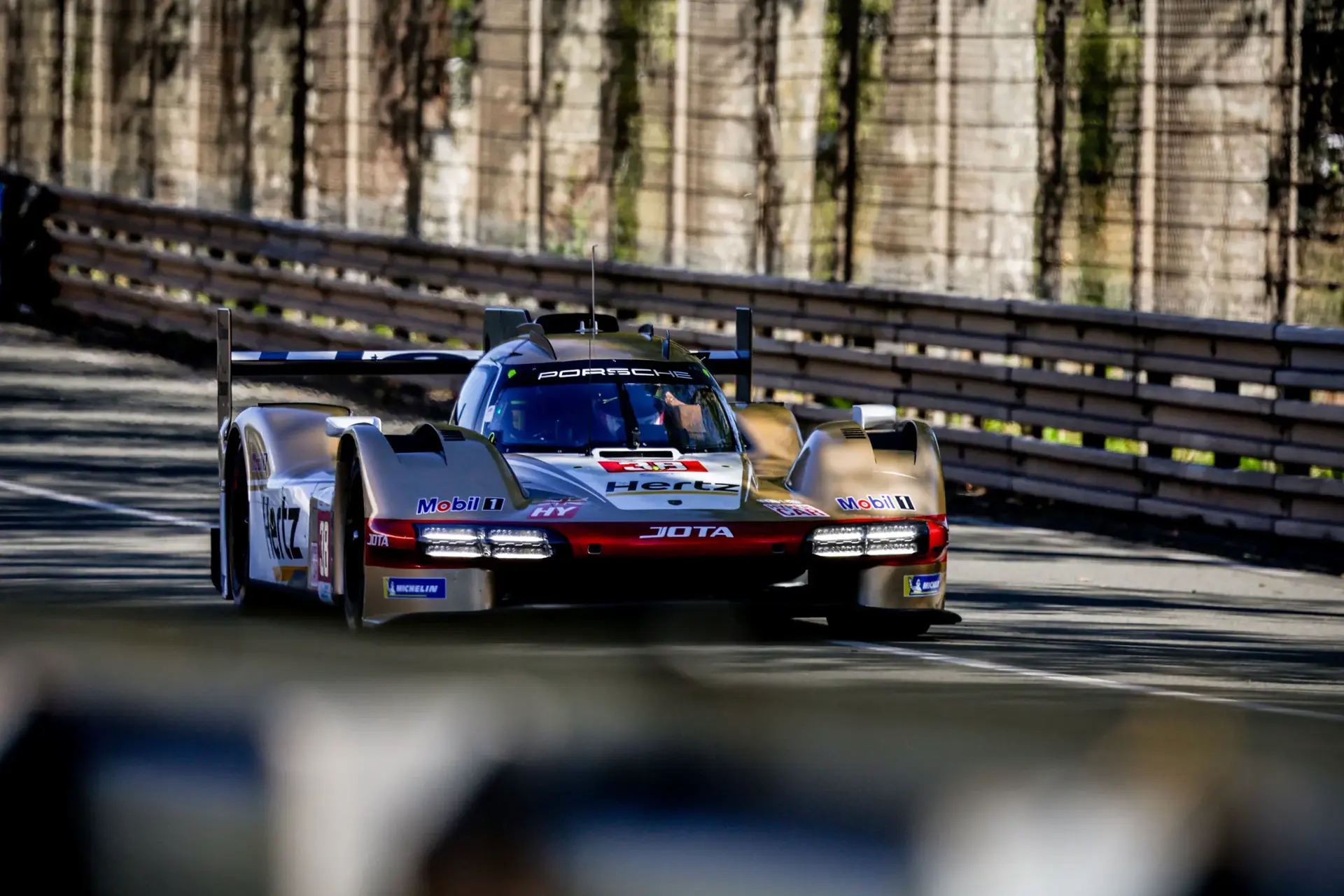 En guld- og rødtonet Porsche-racerbil fra JOTA-teamet kører i høj fart på en afspærret racerbane.