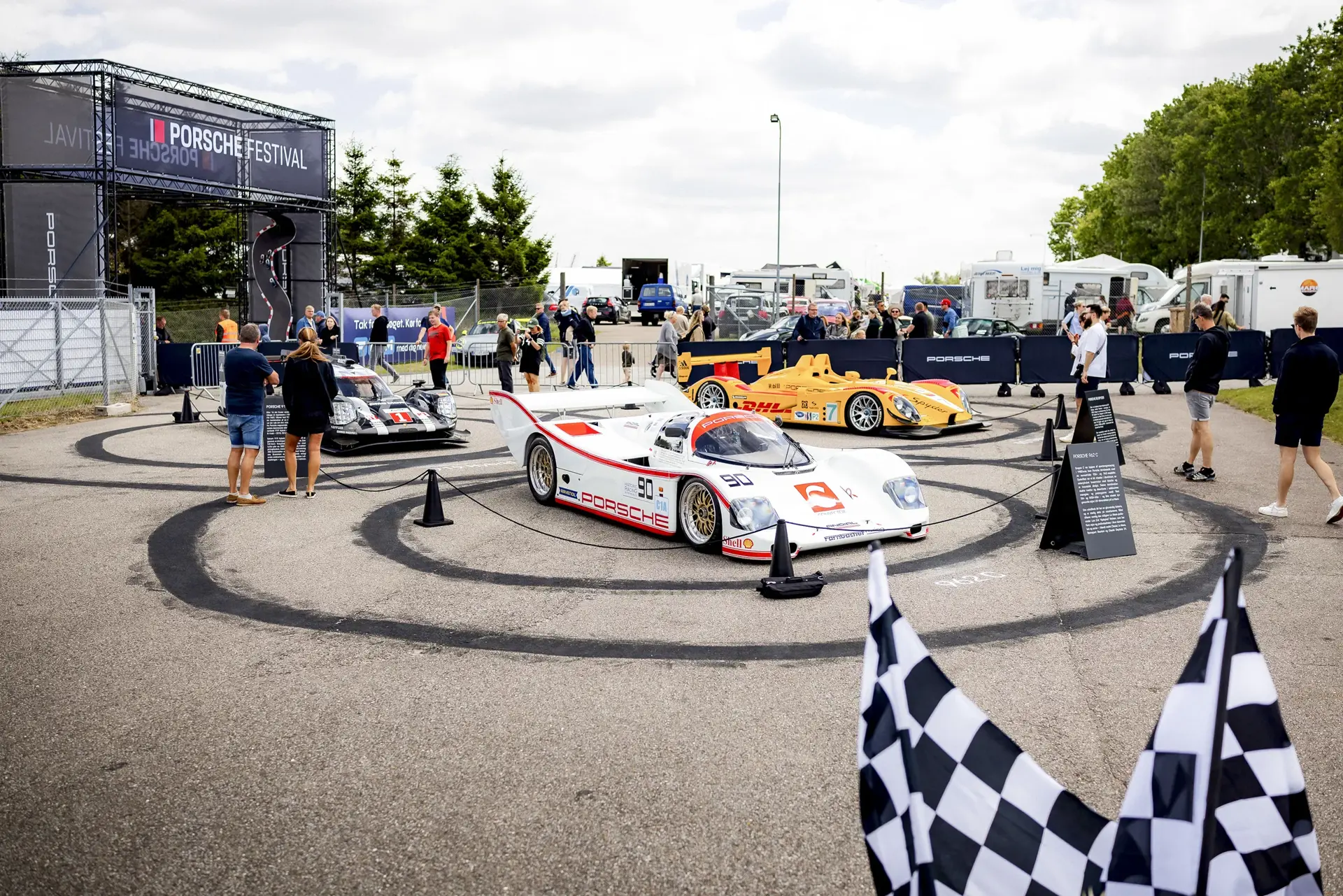 Publikum betragter udstillede Porsche-racerbiler på et udendørs område under Porsche Festival.