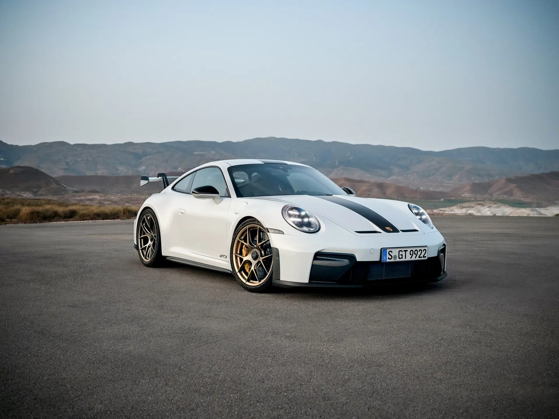Hvid Porsche 911 GT3 med sort stribe og stor hækspoiler holder parkeret på en åben vej med bjerge i baggrunden.