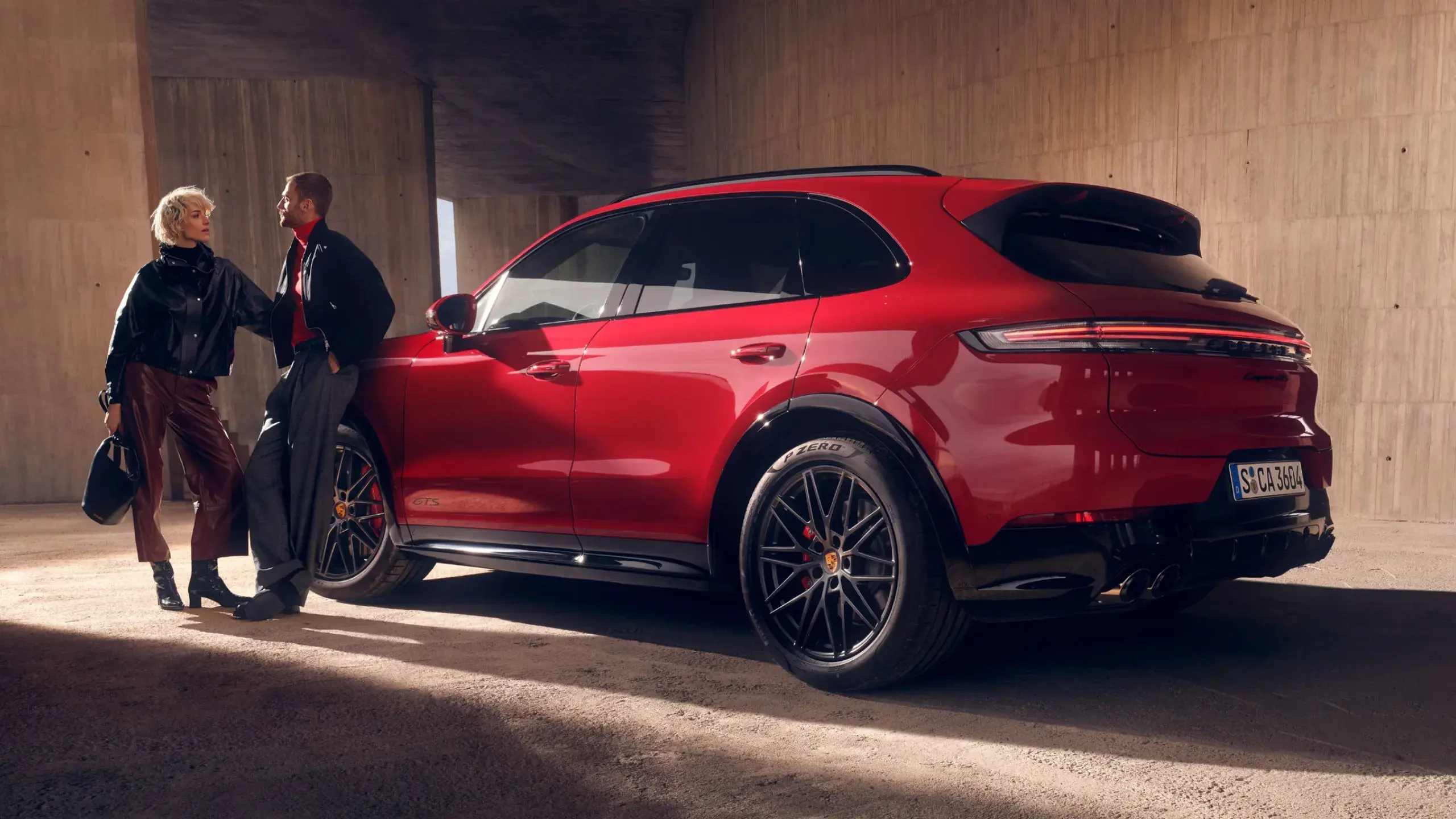 En mand i jakkesæt går forbi bagenden af en rød Porsche Cayenne GTS parkeret ved en moderne bygning.
