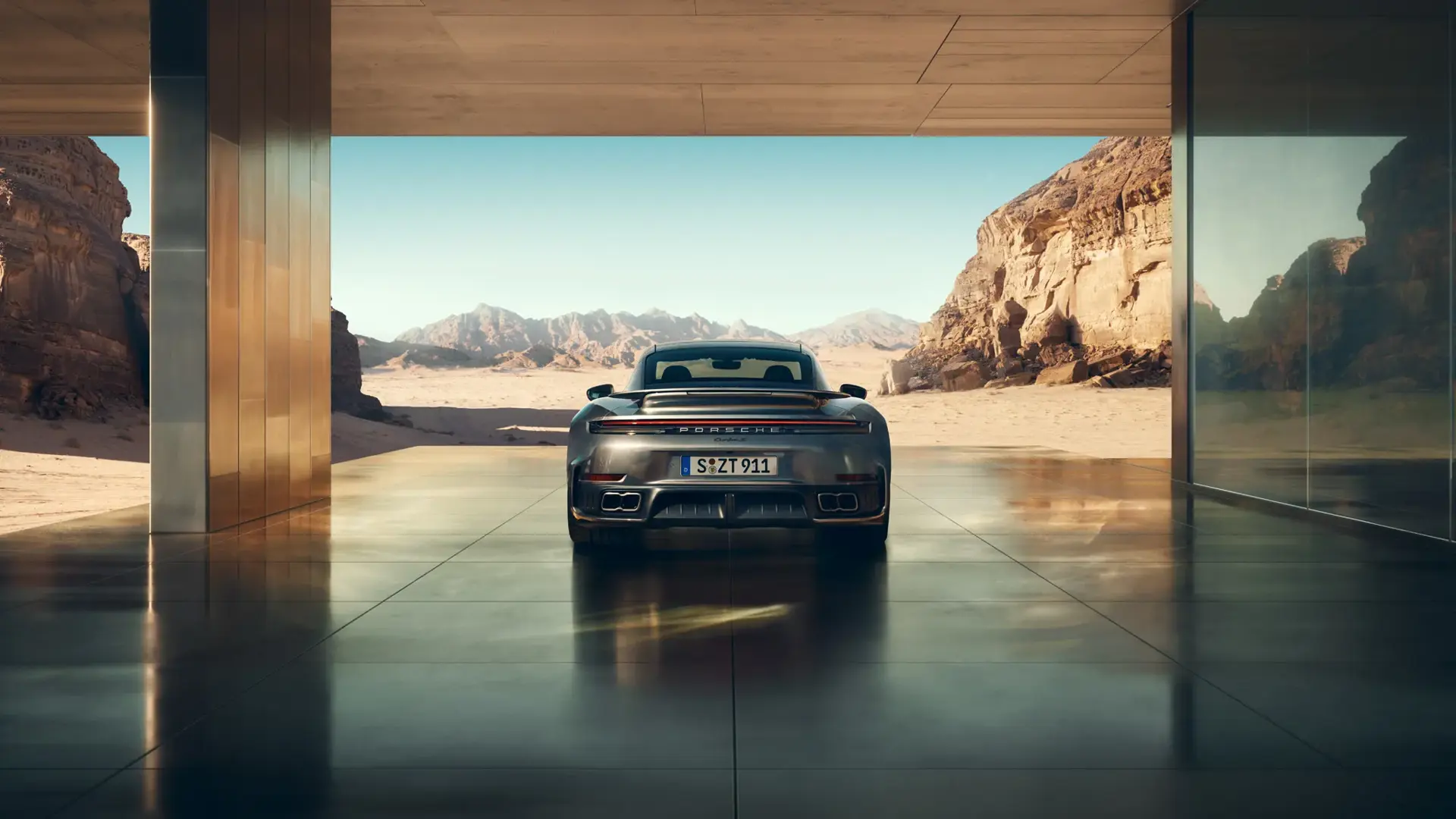 En sølvfarvet Porsche 911 Turbo S set bagfra holder i en moderne, åben bygning med udsigt til en solrig ørken og bjerglandskab.