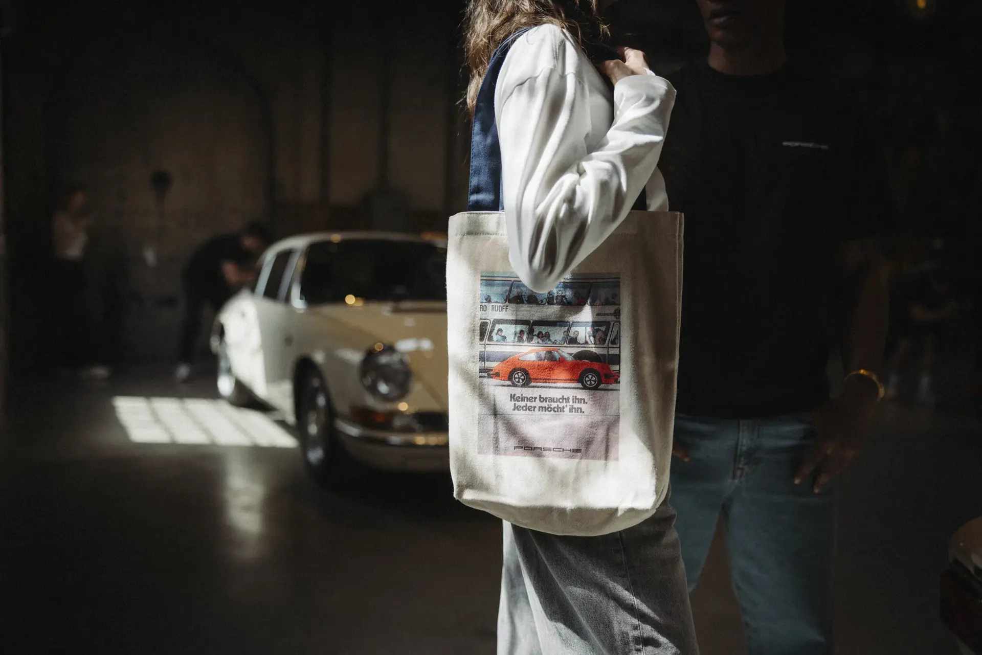 En person bærer en stofpose med et Porsche-motiv foran en klassisk hvid Porsche i en dæmpet, industriel hal.