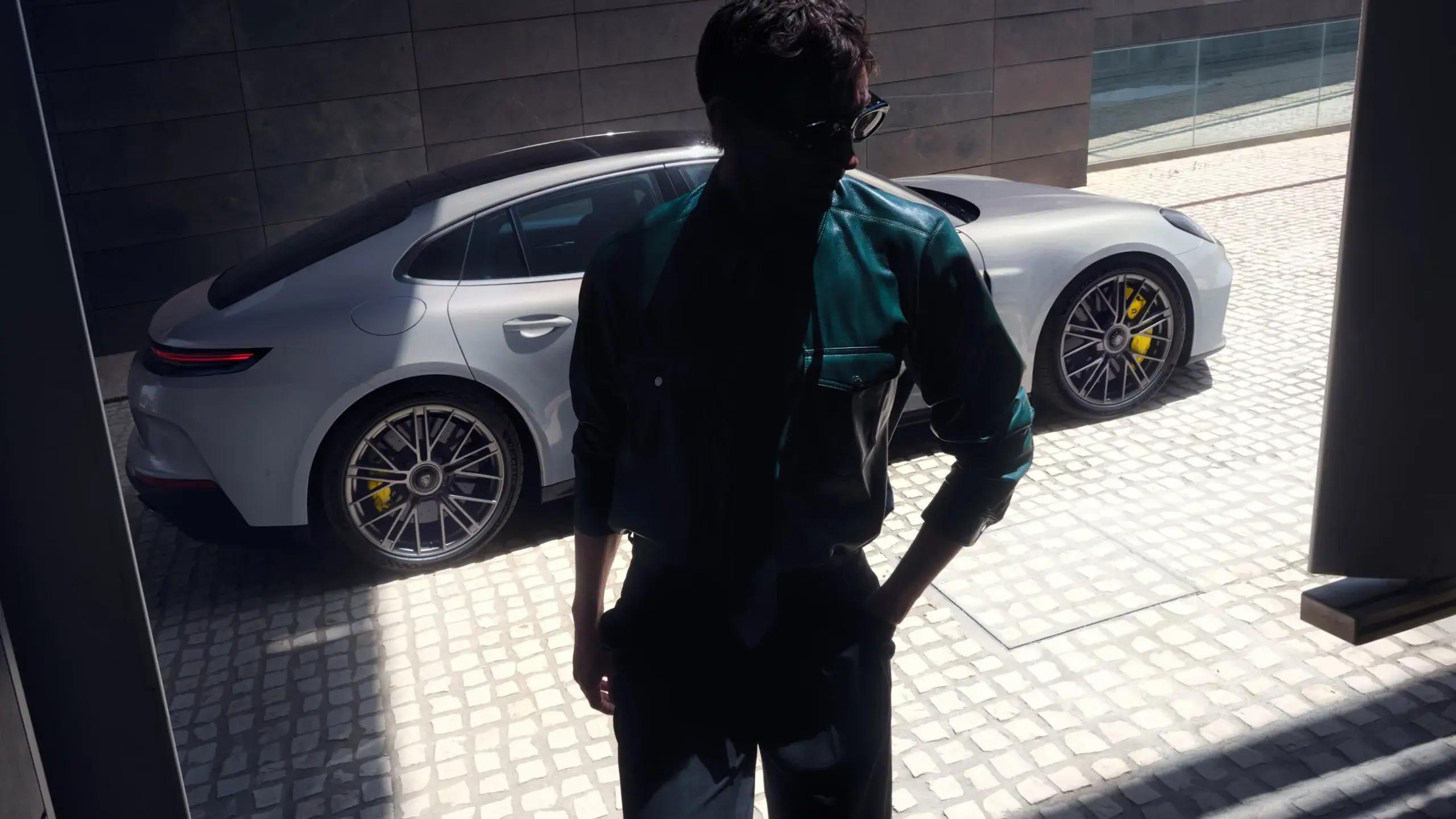 En mand i lys frakke står lænet mod en sølvgrå Porsche Panamera Turbo S E-Hybrid, hvor bilens forhjul med gule bremsekalibre er tydeligt i fokus.