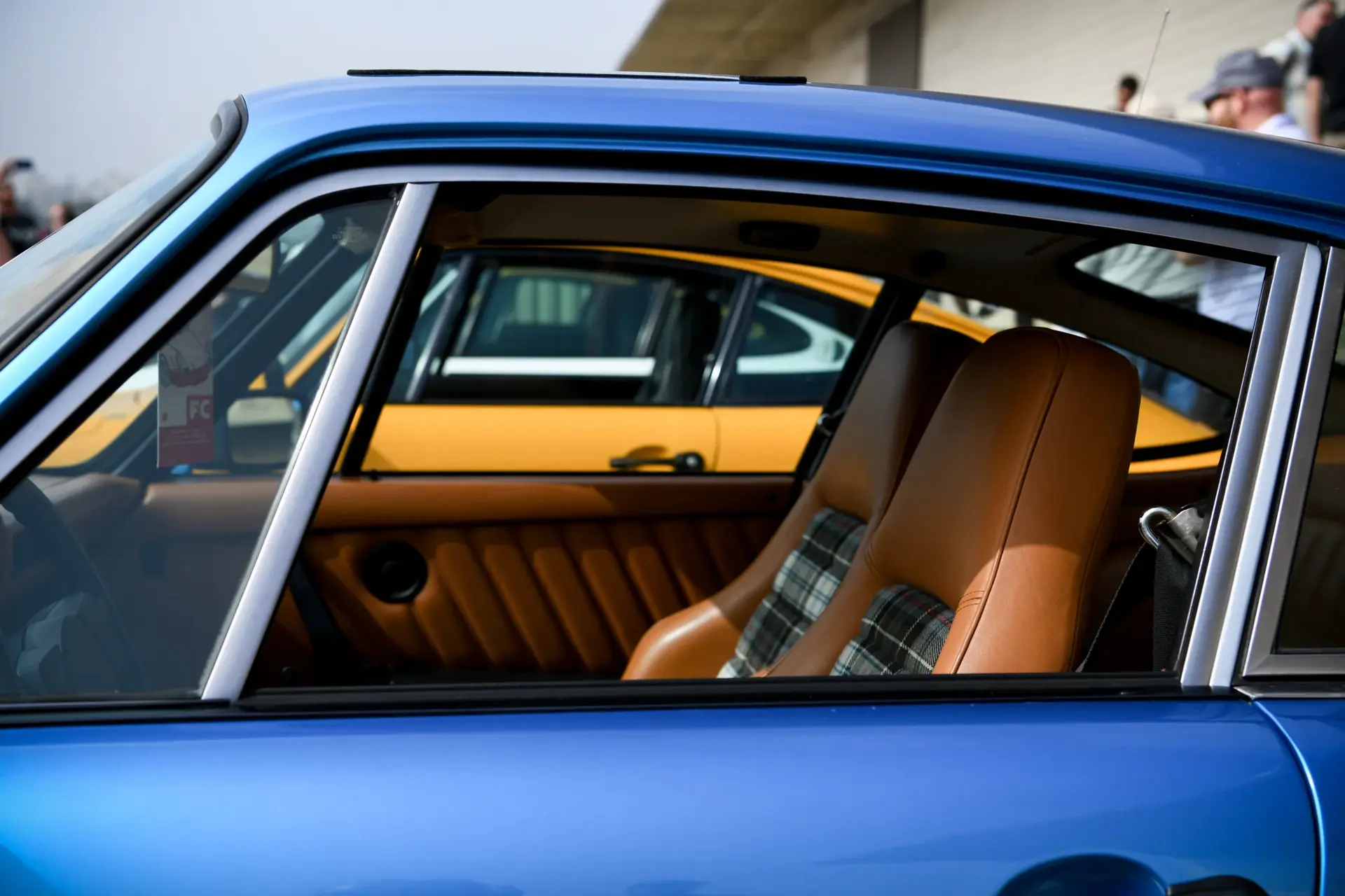 Nærbillede af kabinen i en blå klassisk Porsche med brune sæder og andre farvede 911’ere parkeret ved siden af.
