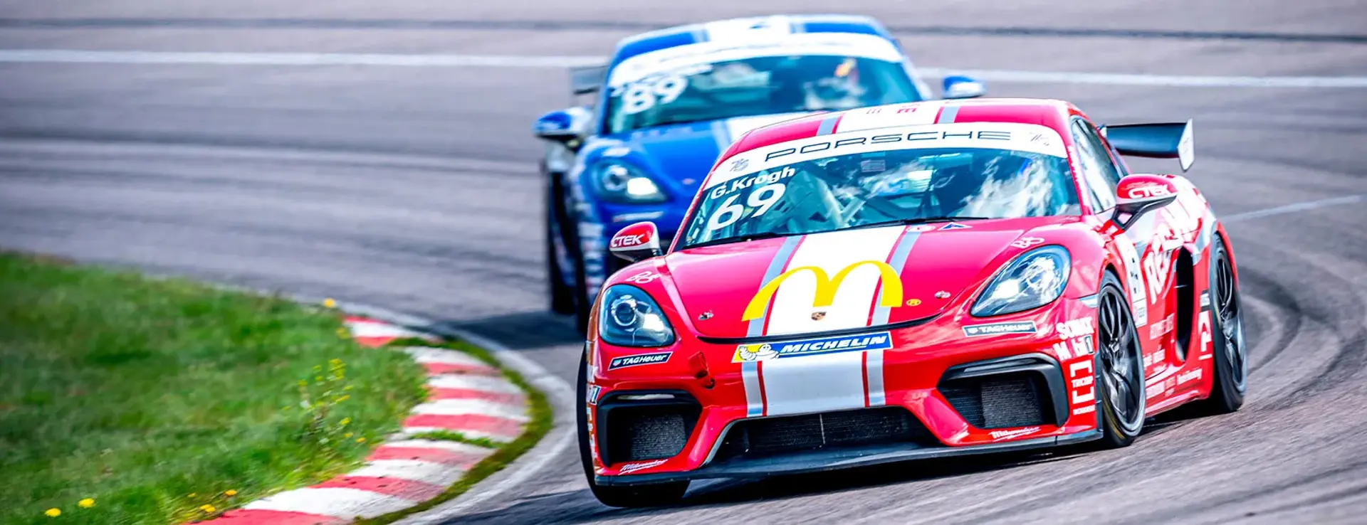 To racerbiler i Porsche-løb tager et sving på Anderstorp-banen, med en rød bil i front og en blå lige bagved.