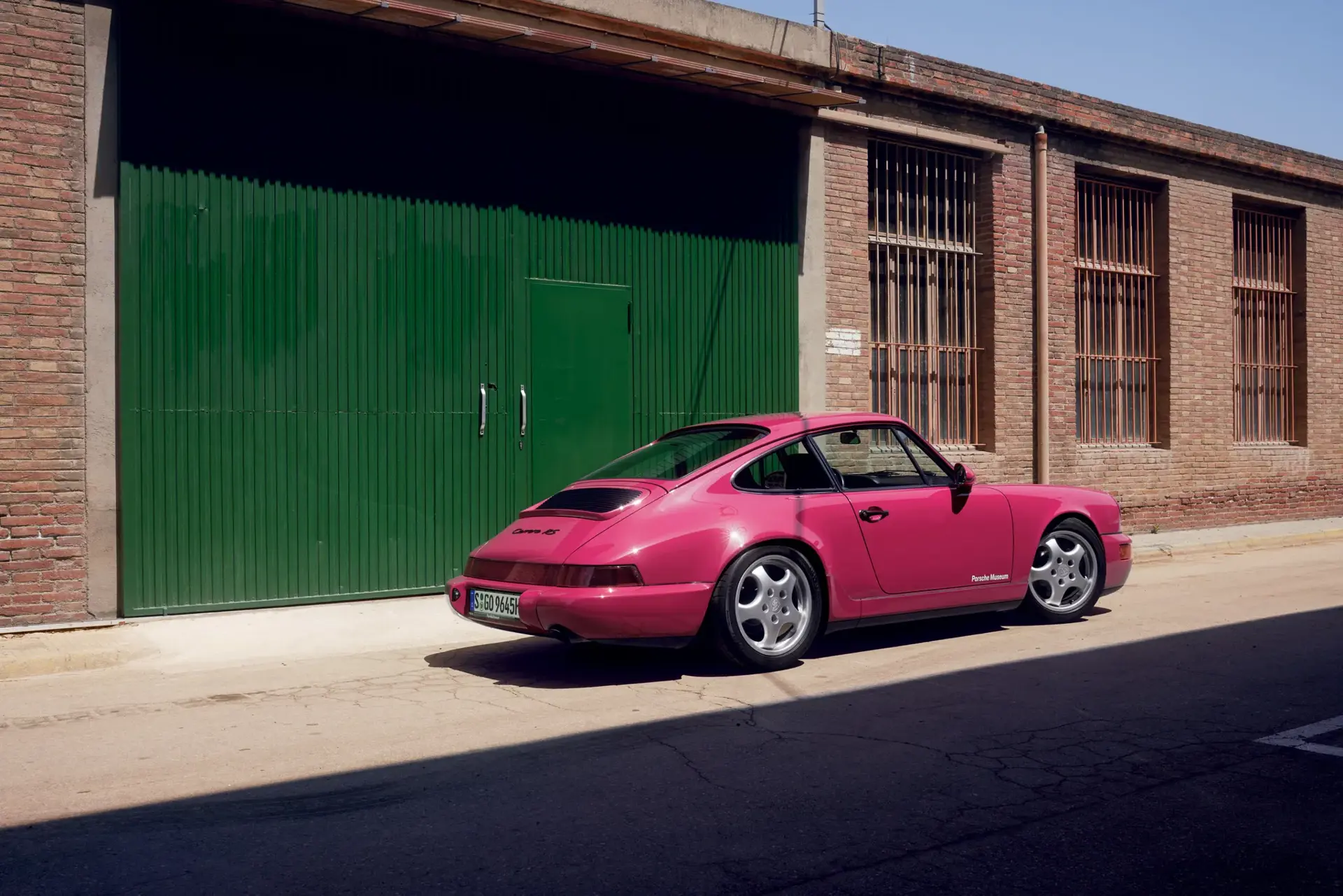 En lyserød klassisk Porsche 911 står parkeret foran en grøn fabriksport ved en murstensbygning.