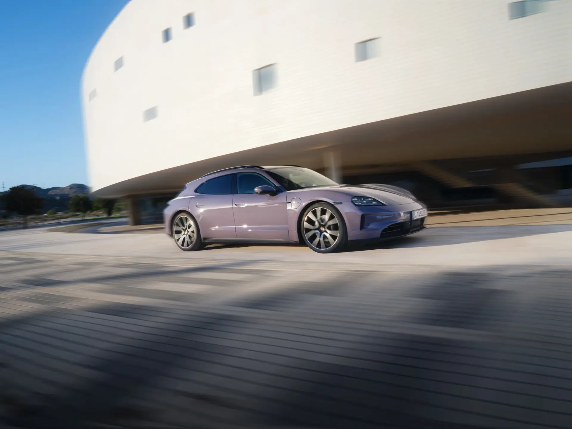 En lilla Porsche Taycan 4S Sport Turismo kører hurtigt forbi en moderne hvid bygning i solskin.