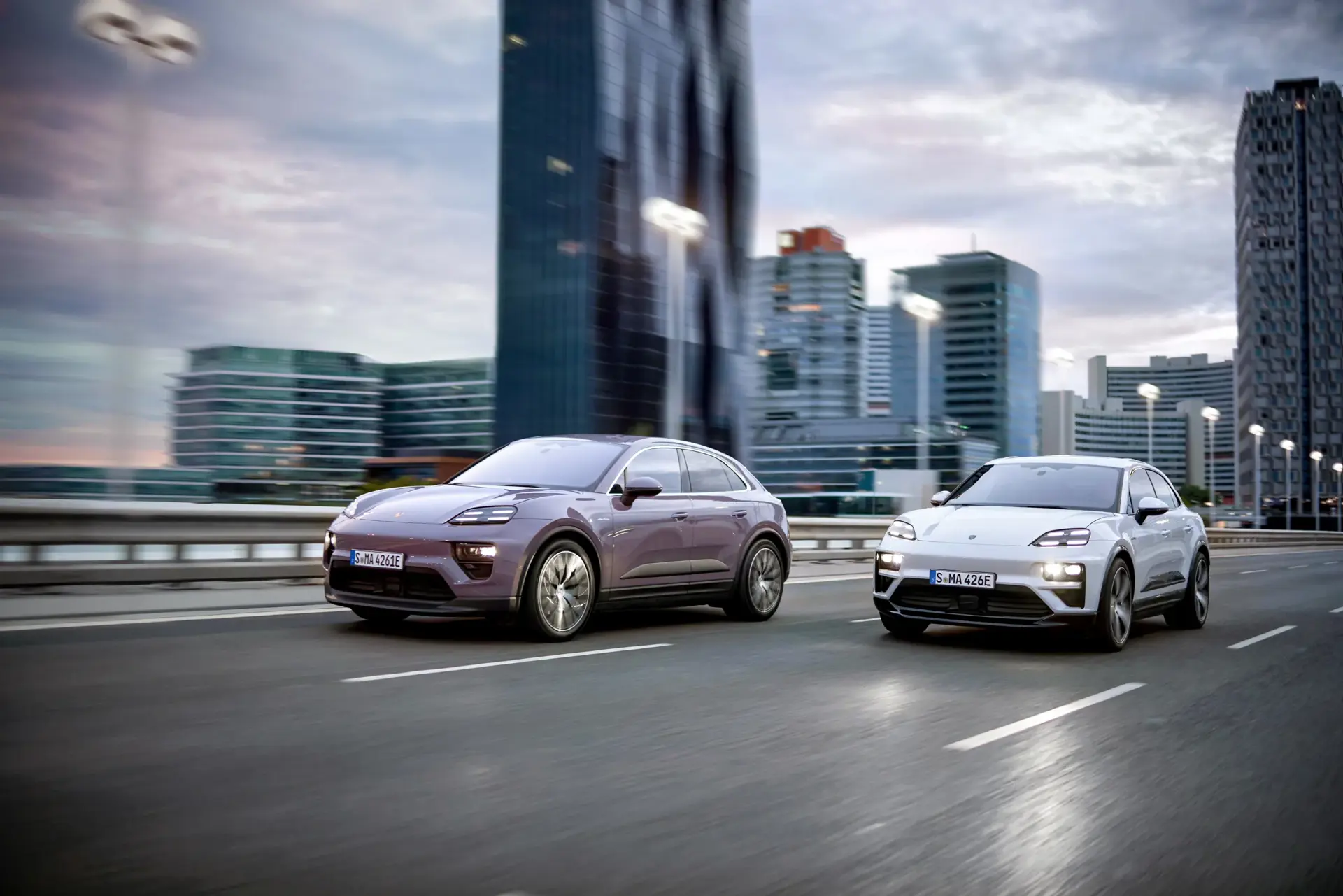 To Porsche Macan elbiler kører side om side på en bymotorvej med moderne højhuse i baggrunden.