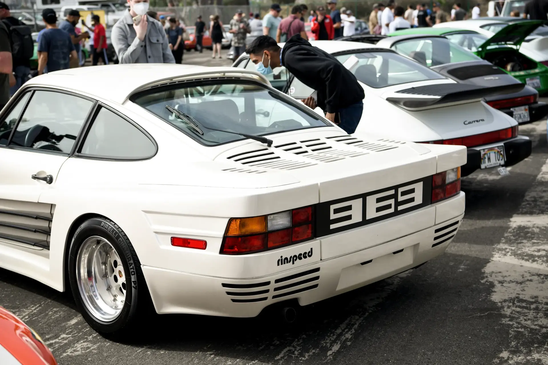 Personer betragter en hvid Rinspeed 969 sportsvogn parkeret blandt andre klassiske Porsche-modeller ved et biltræf.