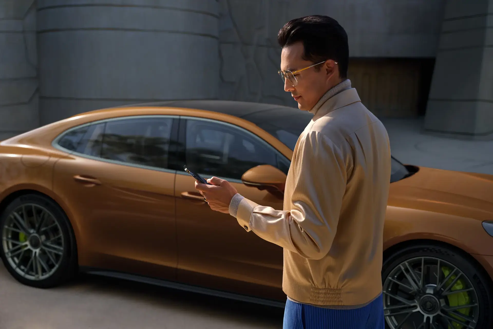 Mand står med sin smartphone foran en gylden Porsche Panamera parkeret i sollys.
