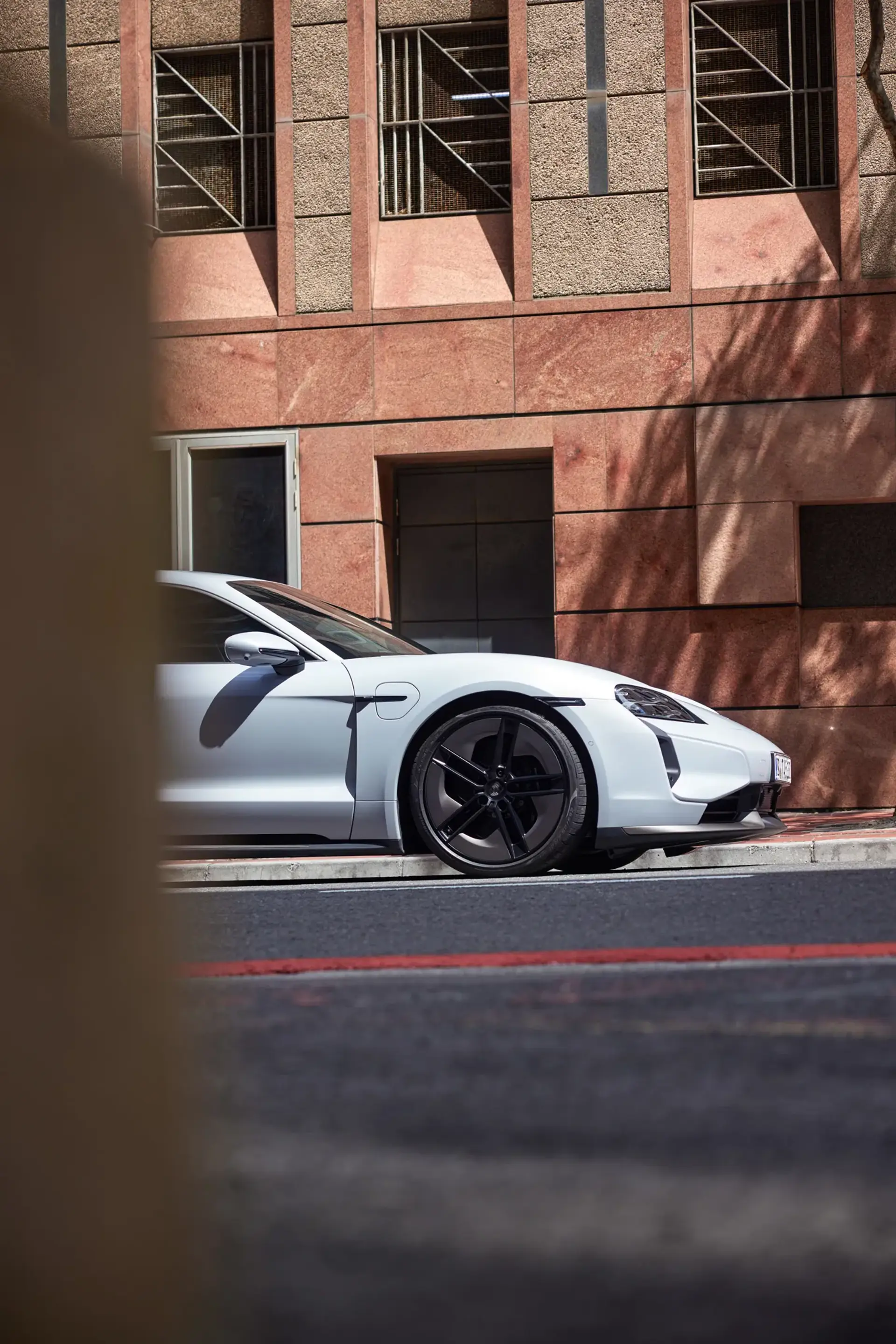 En hvid Porsche Taycan er parkeret ved en kantsten foran en moderne bygning med rødlige sten.