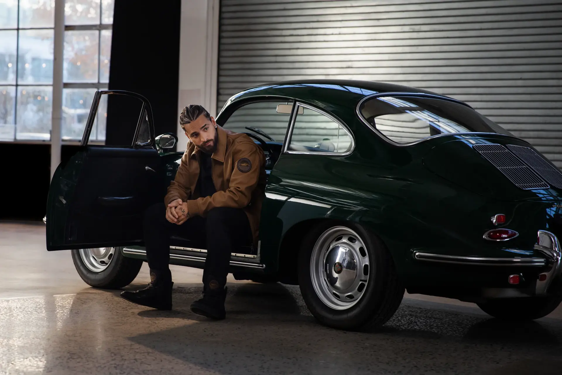 En mand i brun jakke sidder i døren på en mørkegrøn klassisk Porsche parkeret i en industriel hal.