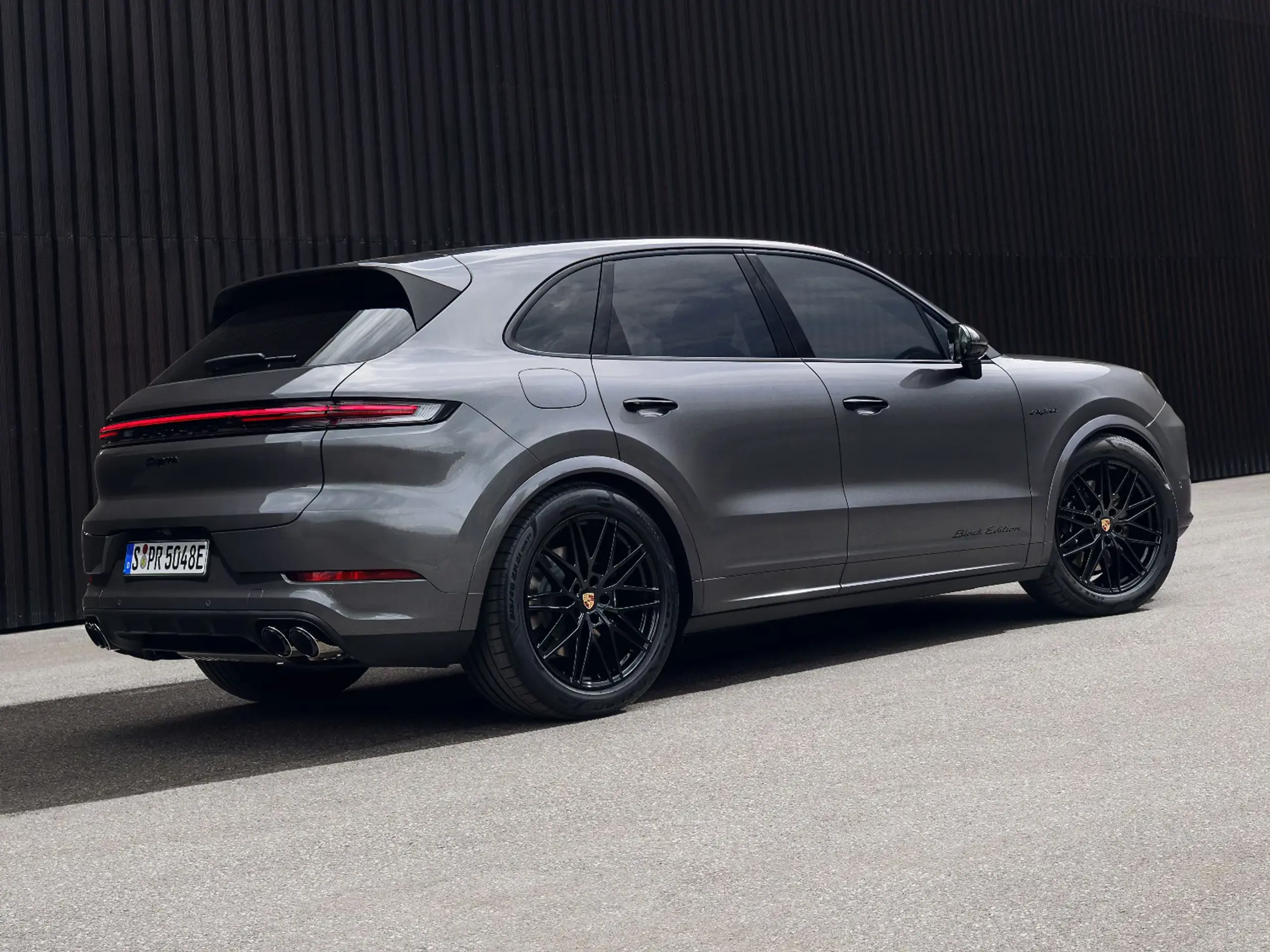 Grå Porsche Cayenne Black Edition SUV set skråt bagfra parkeret foran en mørk væg.