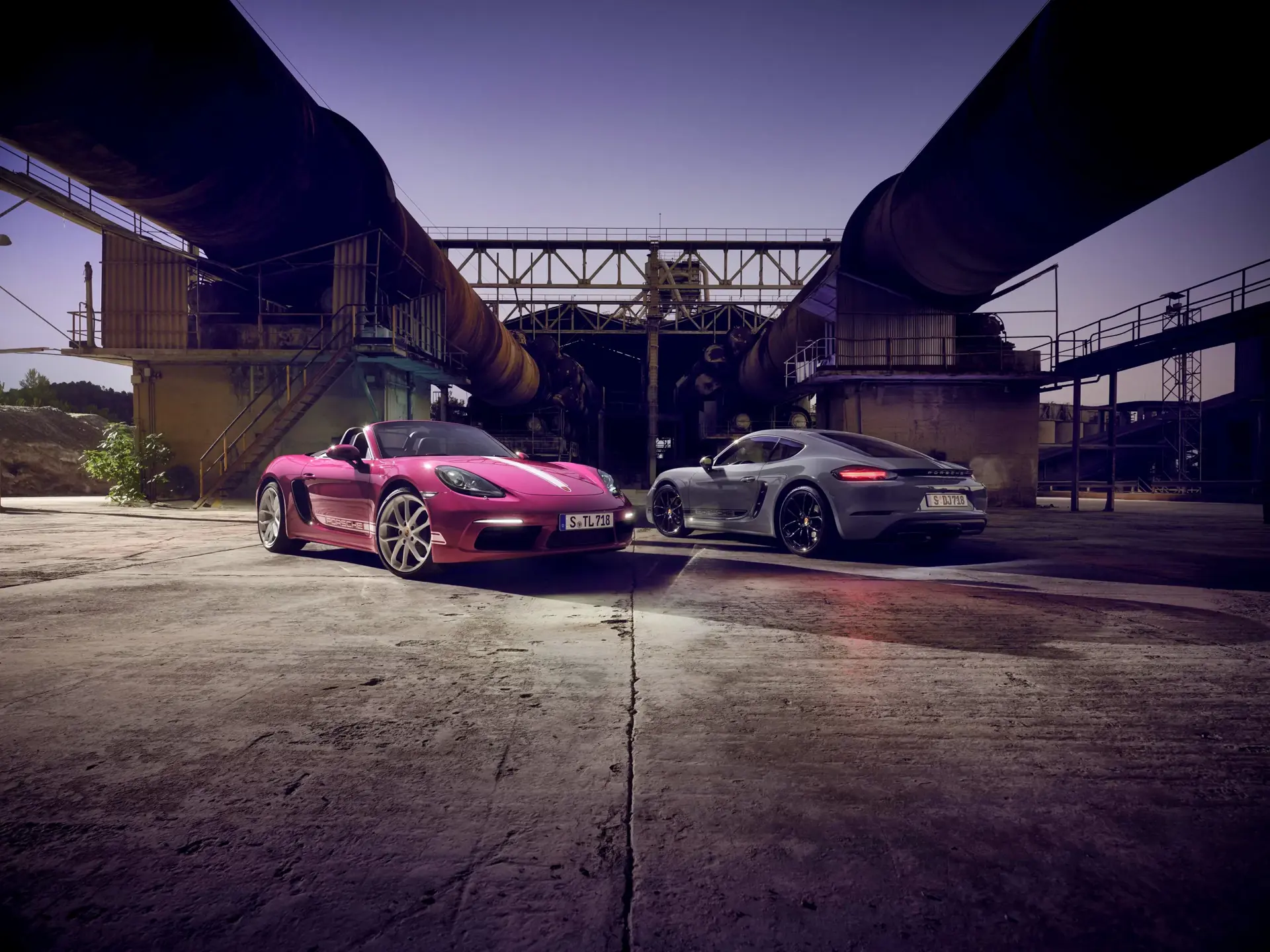 To sportsvogne, en pink cabriolet og en sølvgrå coupé, står parkeret foran et industrielt anlæg i aftenslys.