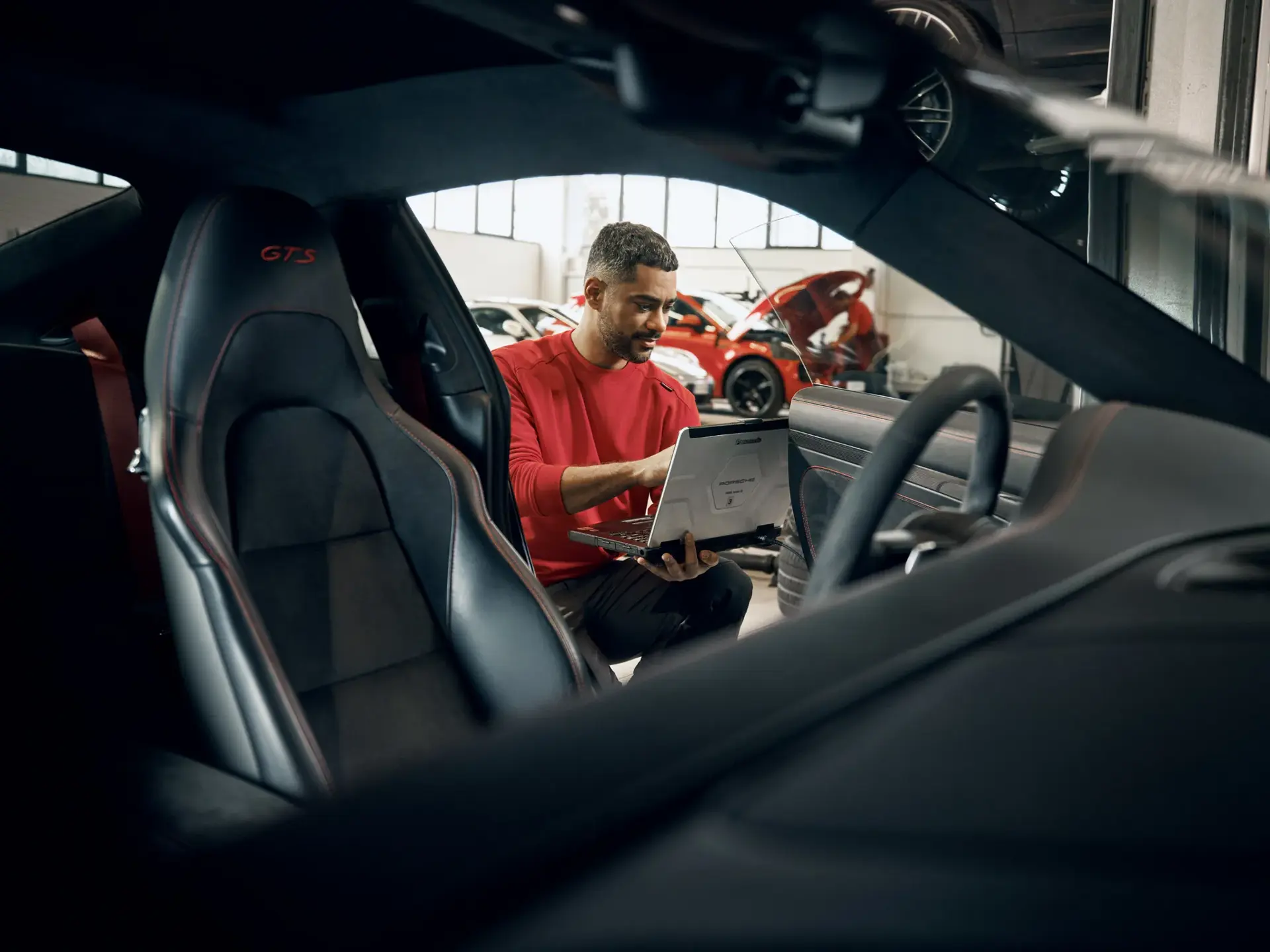Mekaniker i rødt arbejdstøj sidder ved en Porsche og foretager diagnostics på en bærbar computer i et autoværksted.