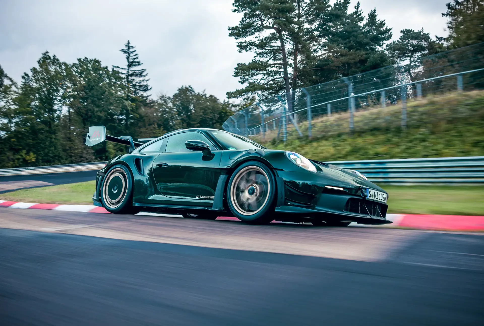 En mørkegrøn Porsche 911 med Manthey-udstyr kører i høj fart på en racerbane omgivet af træer.