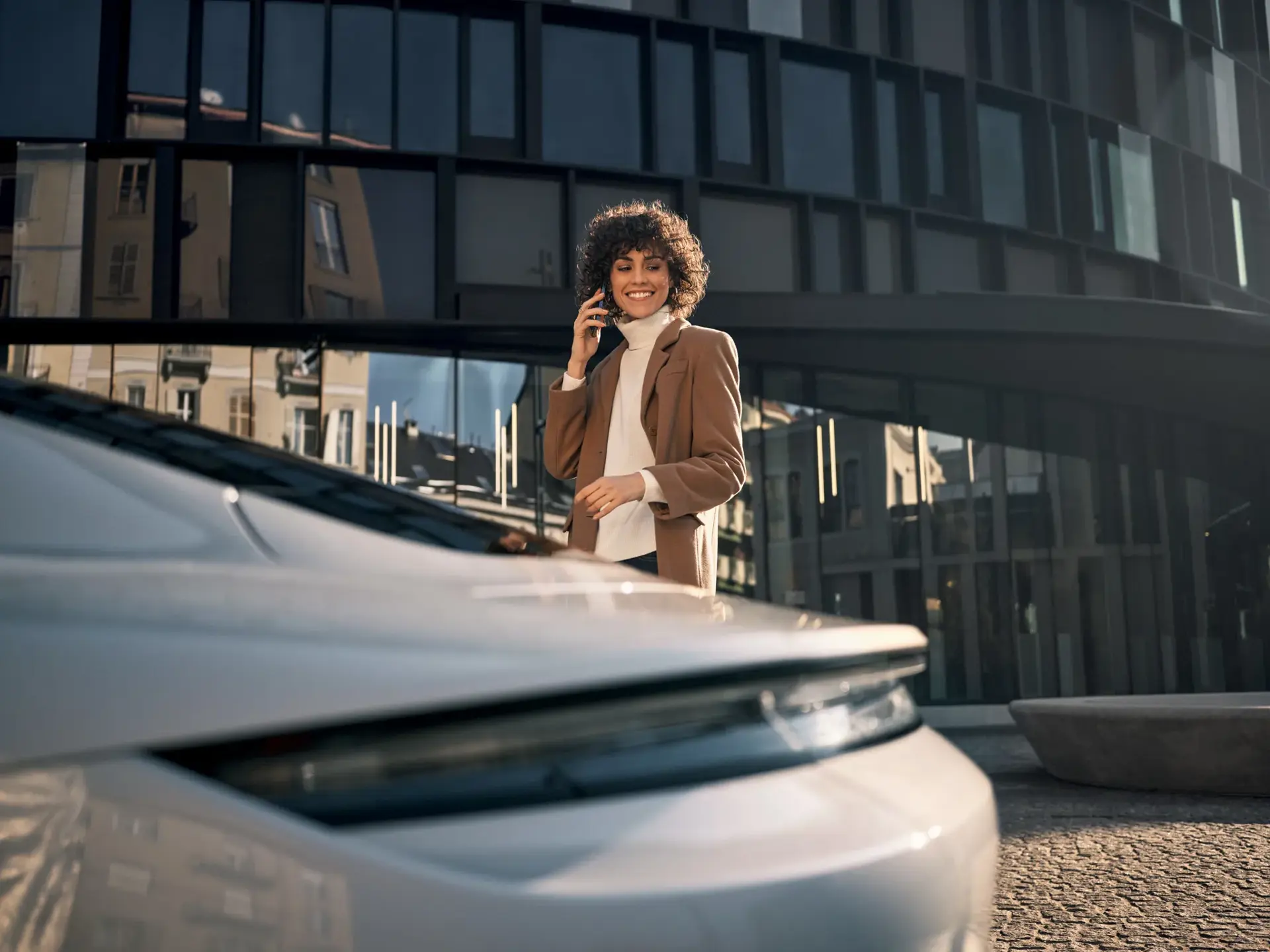 En smilende person står ved en hvid Porsche i byen og taler i telefon foran en moderne bygning.