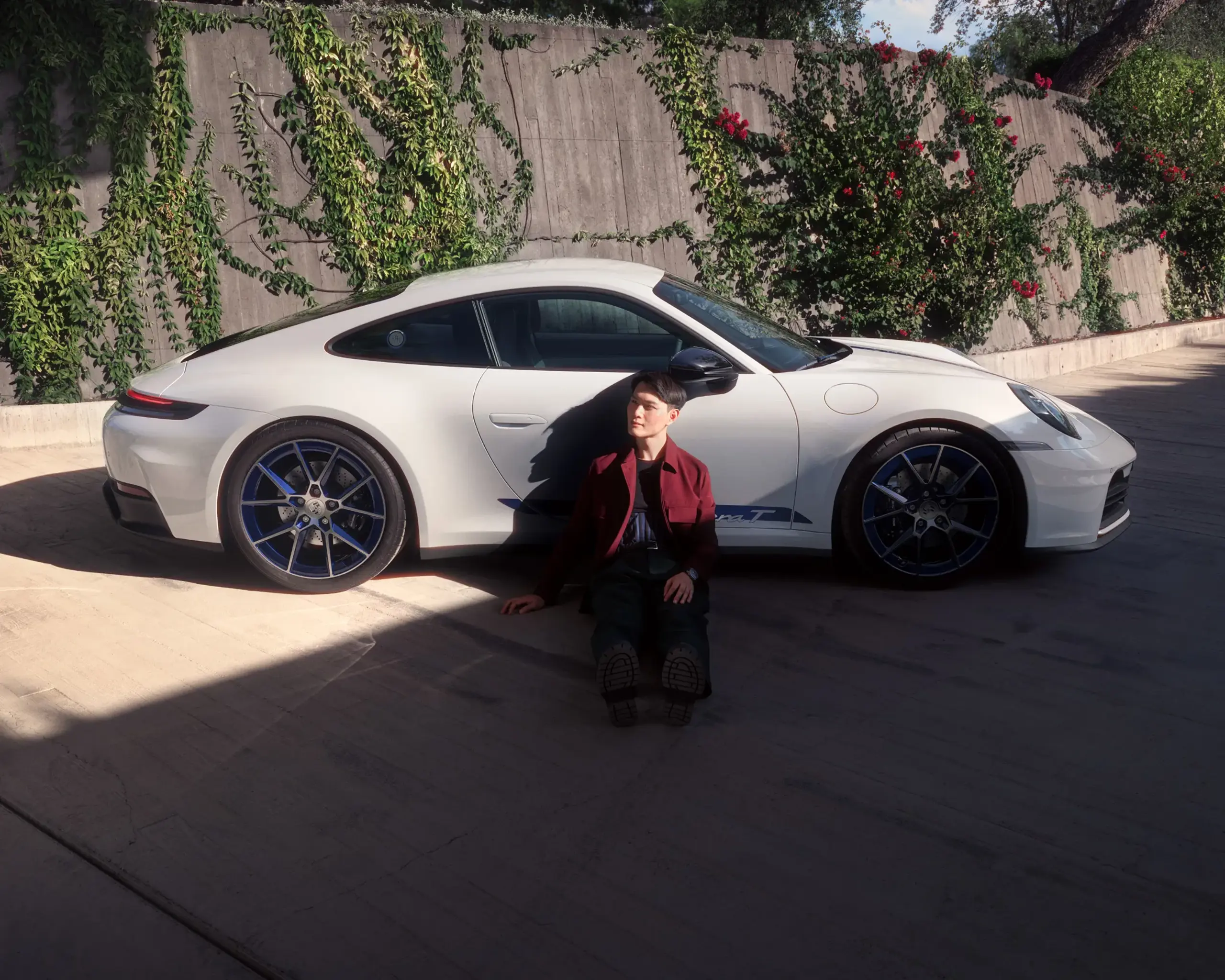 En person sidder på jorden foran en hvid Porsche 911 Carrera T parkeret ved en mur med grønne klatreplanter.