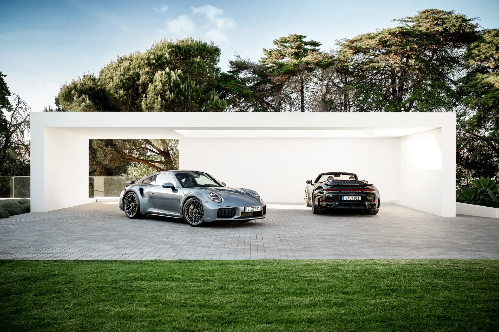 To Porsche 911 sportsvogne holder parkeret foran en moderne, hvid carport omgivet af grønne træer.
