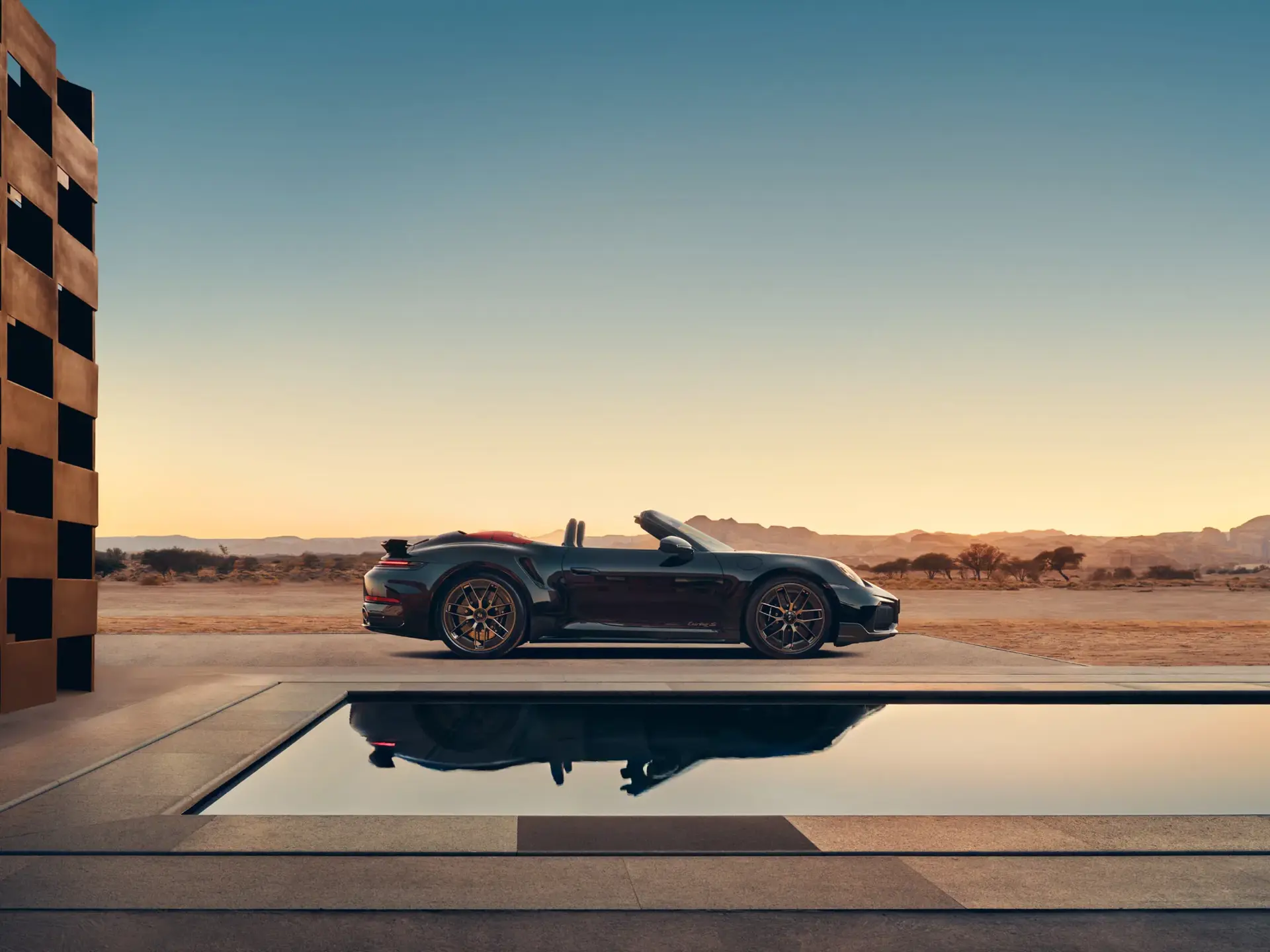 En sort Porsche 911 Turbo S cabriolet holder parkeret ved en moderne pool i ørkenlandskab ved solnedgang.