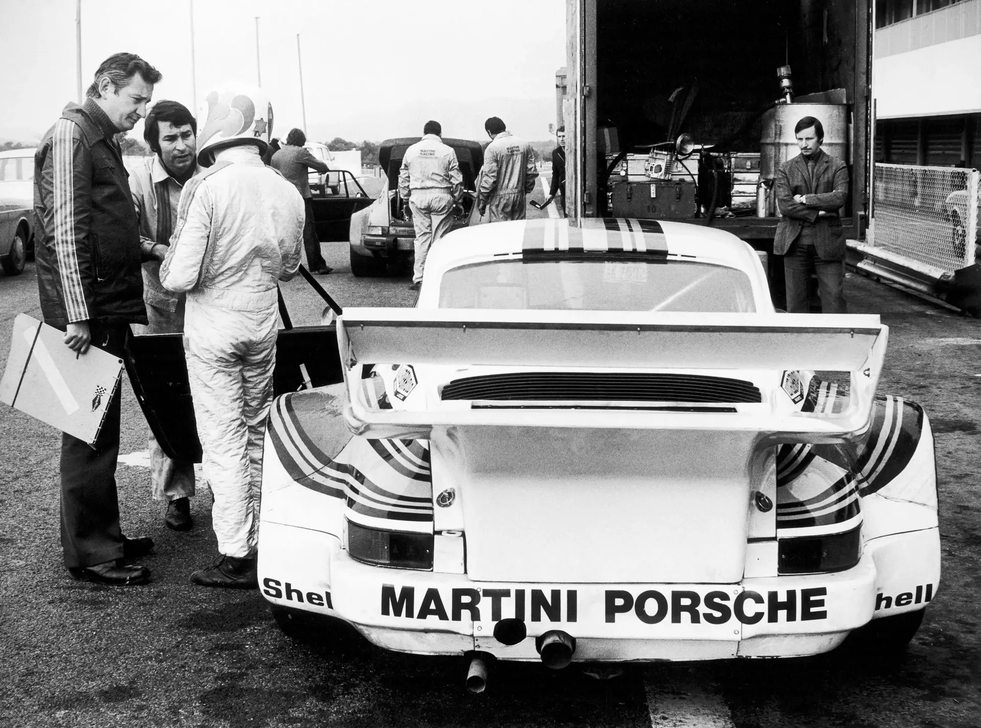 Sort-hvidt foto af en MARTINI PORSCHE-racerbil i pitområdet, omgivet af mekanikere og kørere under testkørslerne på Paul Ricard-banen.