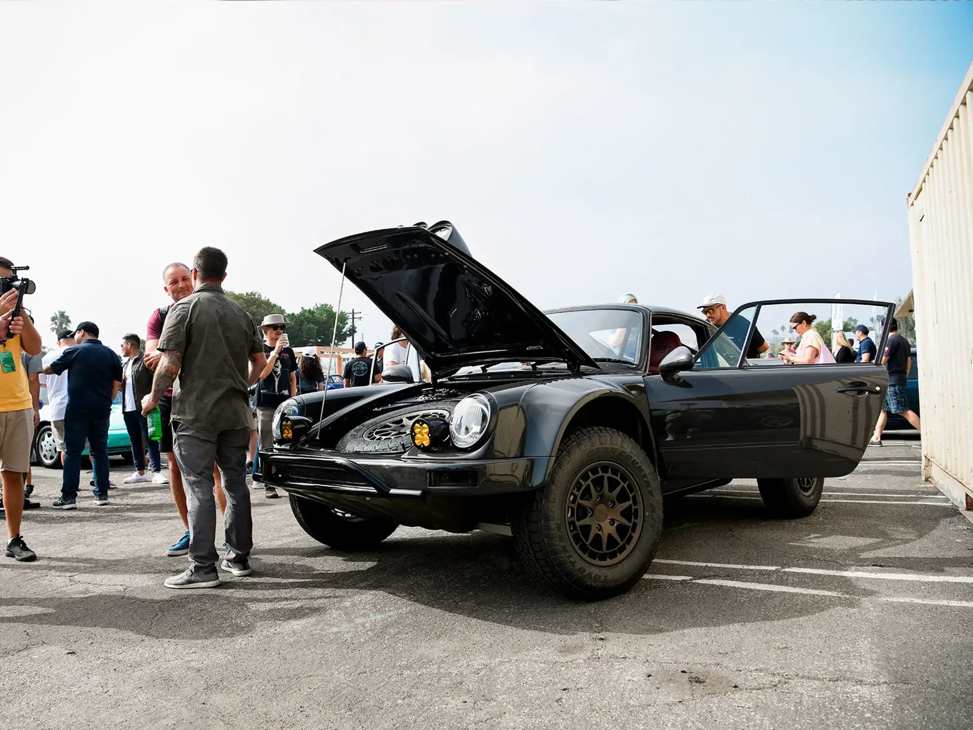 En sort klassisk bil med åben motorhjelm og dør omgivet af mennesker ved et biltræf.
