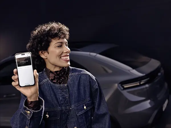 Smilende person foran en bil holder en smartphone op med Porsche Connect digital nøgle på skærmen.