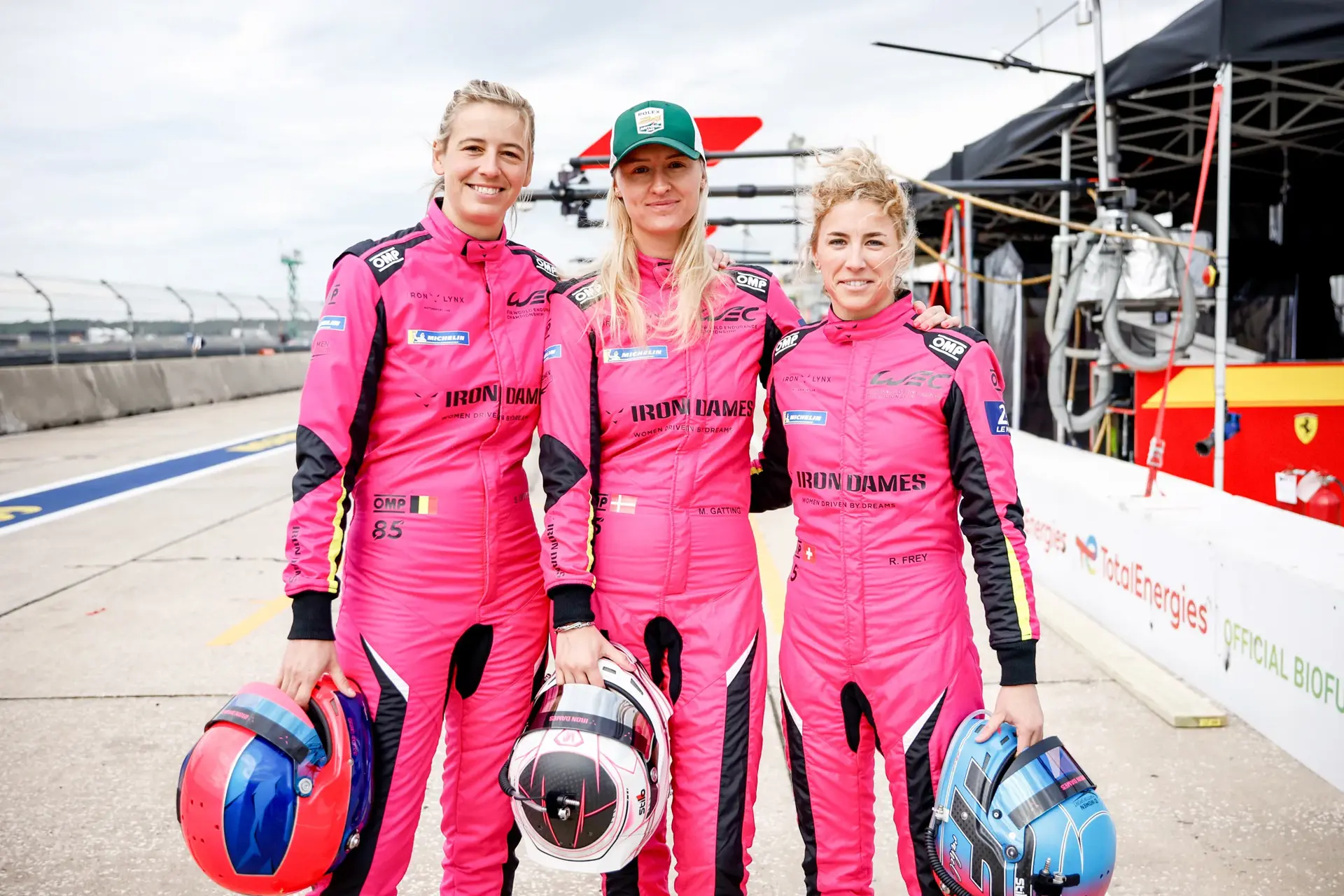 Tre racerkørere i pink Iron Dames-dragter står sammen i pitten og holder deres hjelme.