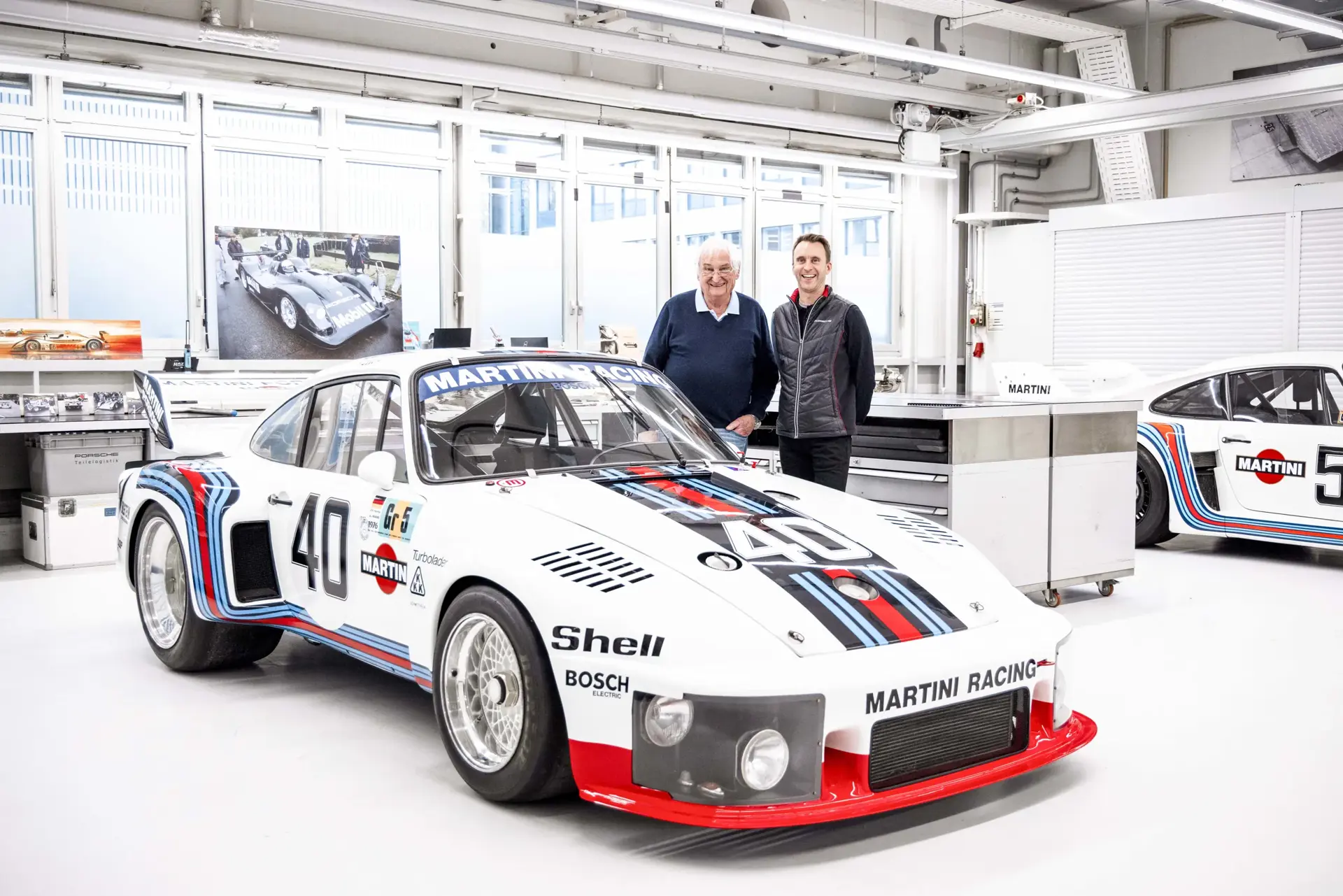 To mænd står i et lyst værksted bag en klassisk hvid Porsche racerbil med Martini Racing-bemaling og startnummer 40.