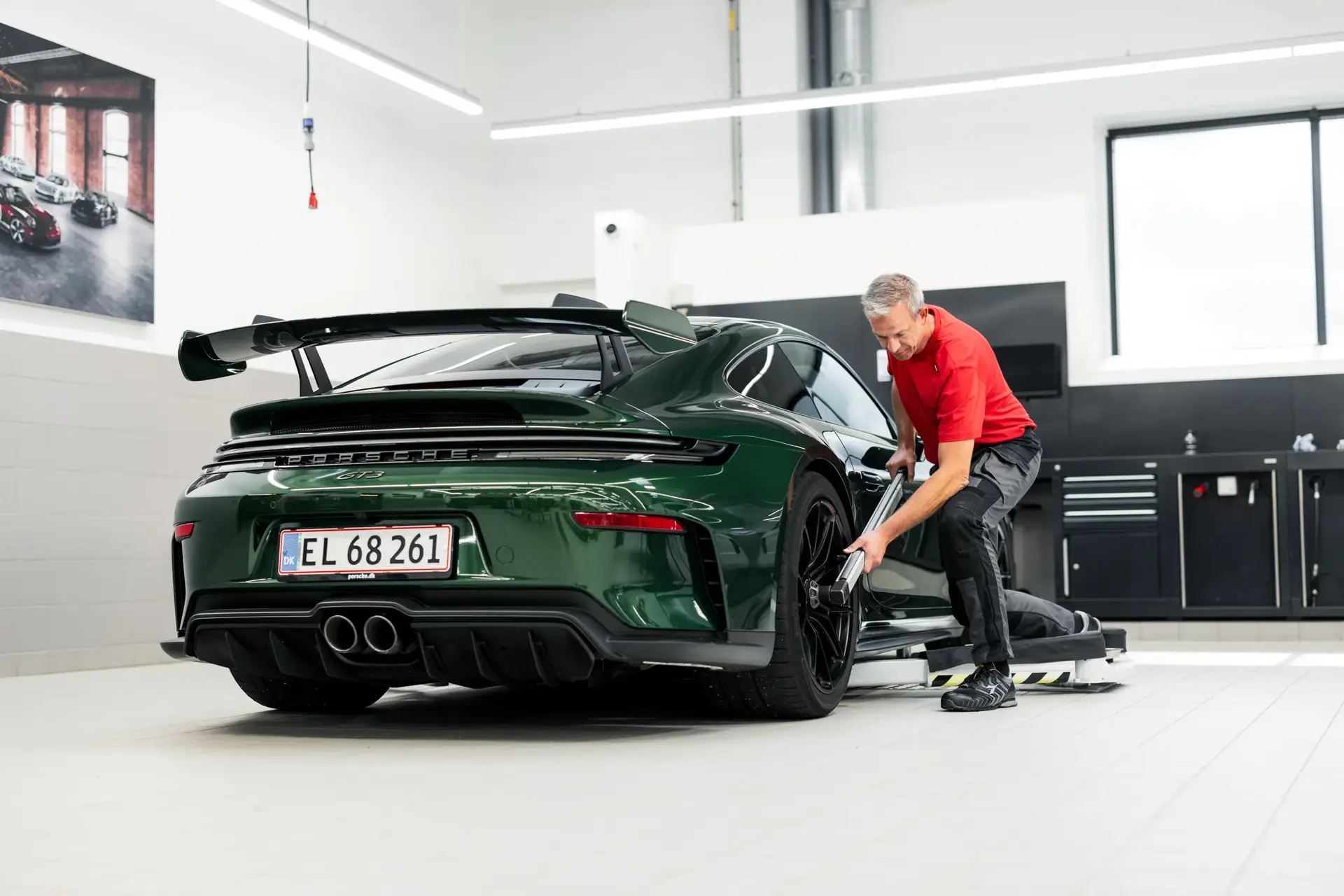 En mekaniker arbejder på et hjul på en mørkegrøn Porsche sportsvogn i et lyst og moderne værksted.