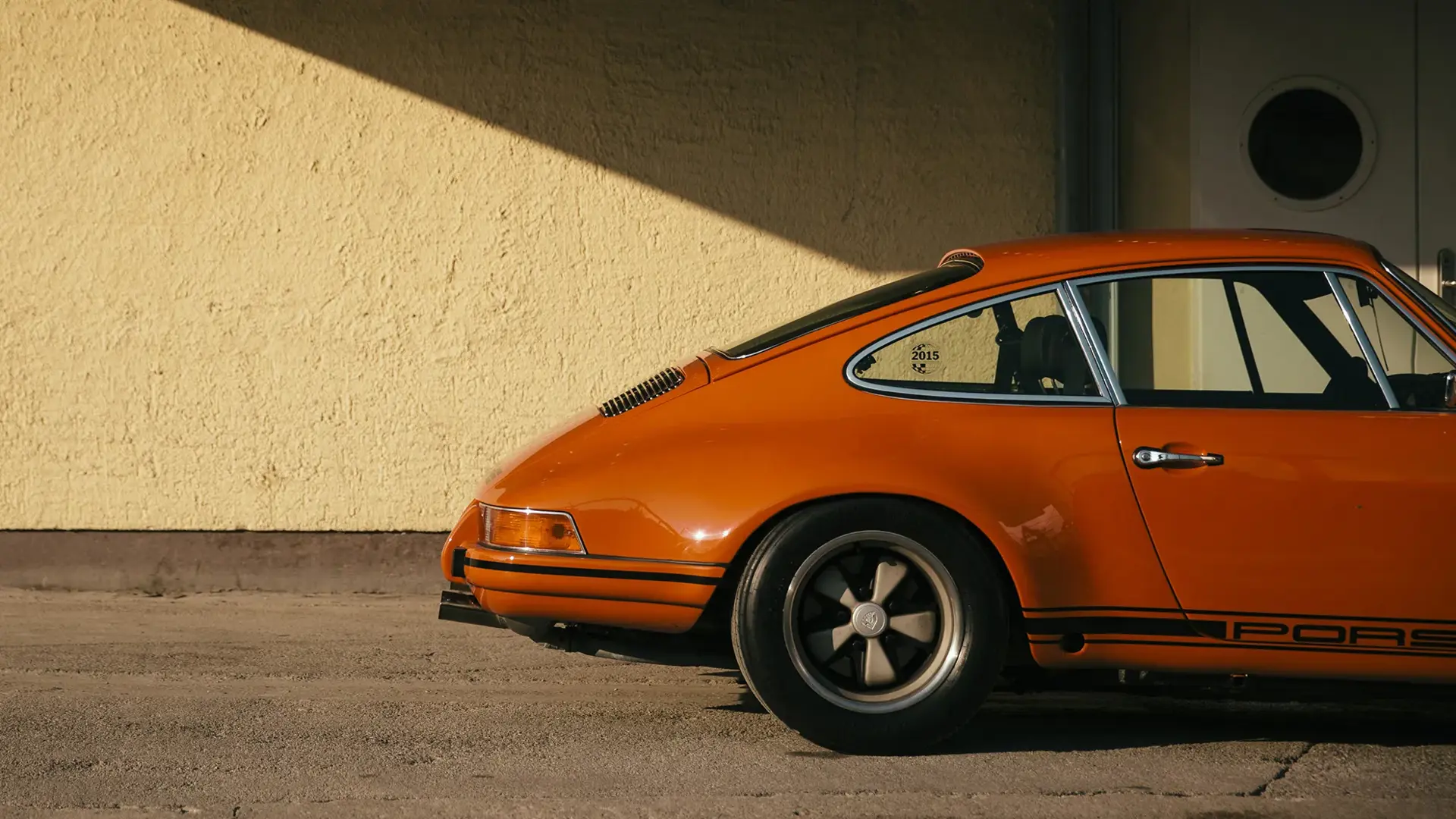 Sidevisning af en orange klassisk sportsvogn parkeret foran en gul mur i sollys.