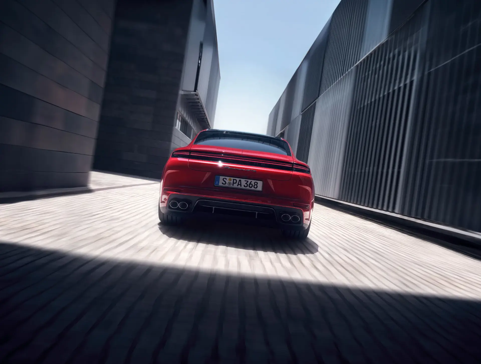 Rød Porsche Panamera GTS set bagfra, der kører hurtigt gennem en smal, moderne bypassage mellem høje bygninger.