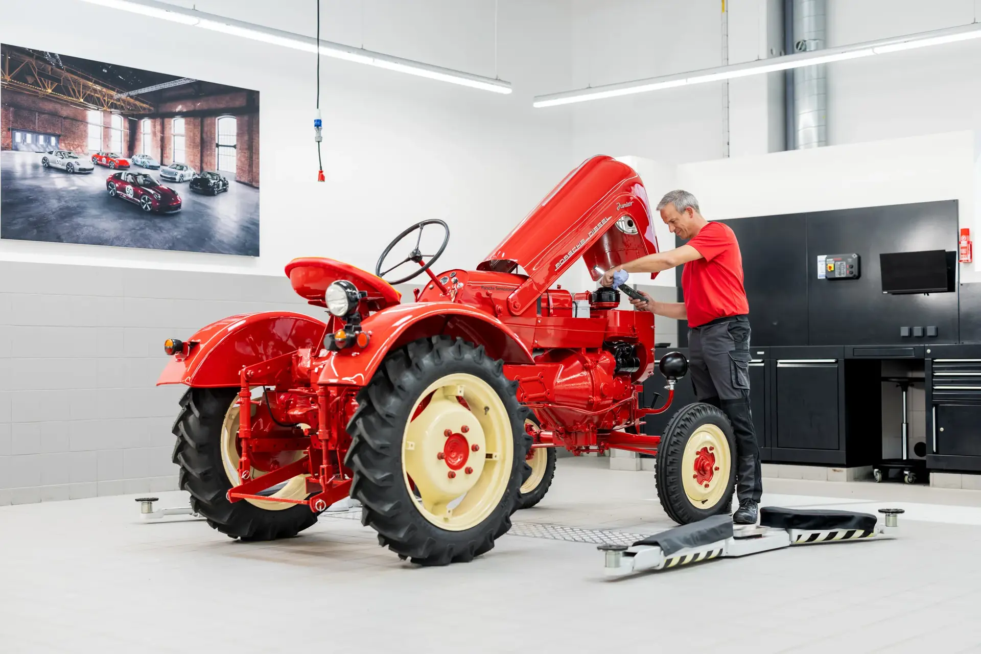 Mekaniker arbejder på en rød klassisk Porsche-traktor i et lyst, moderne værksted.