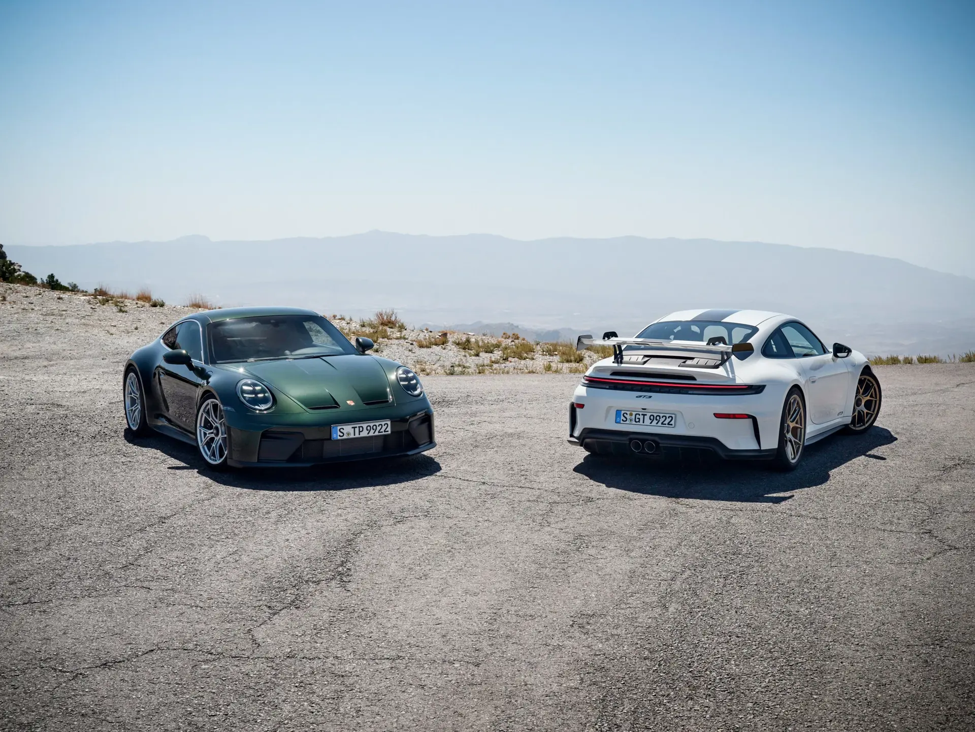 To Porsche 911 GT3 sportsvogne, en grøn og en hvid, holder parkeret på en bjergvej med udsigt over et diset landskab.