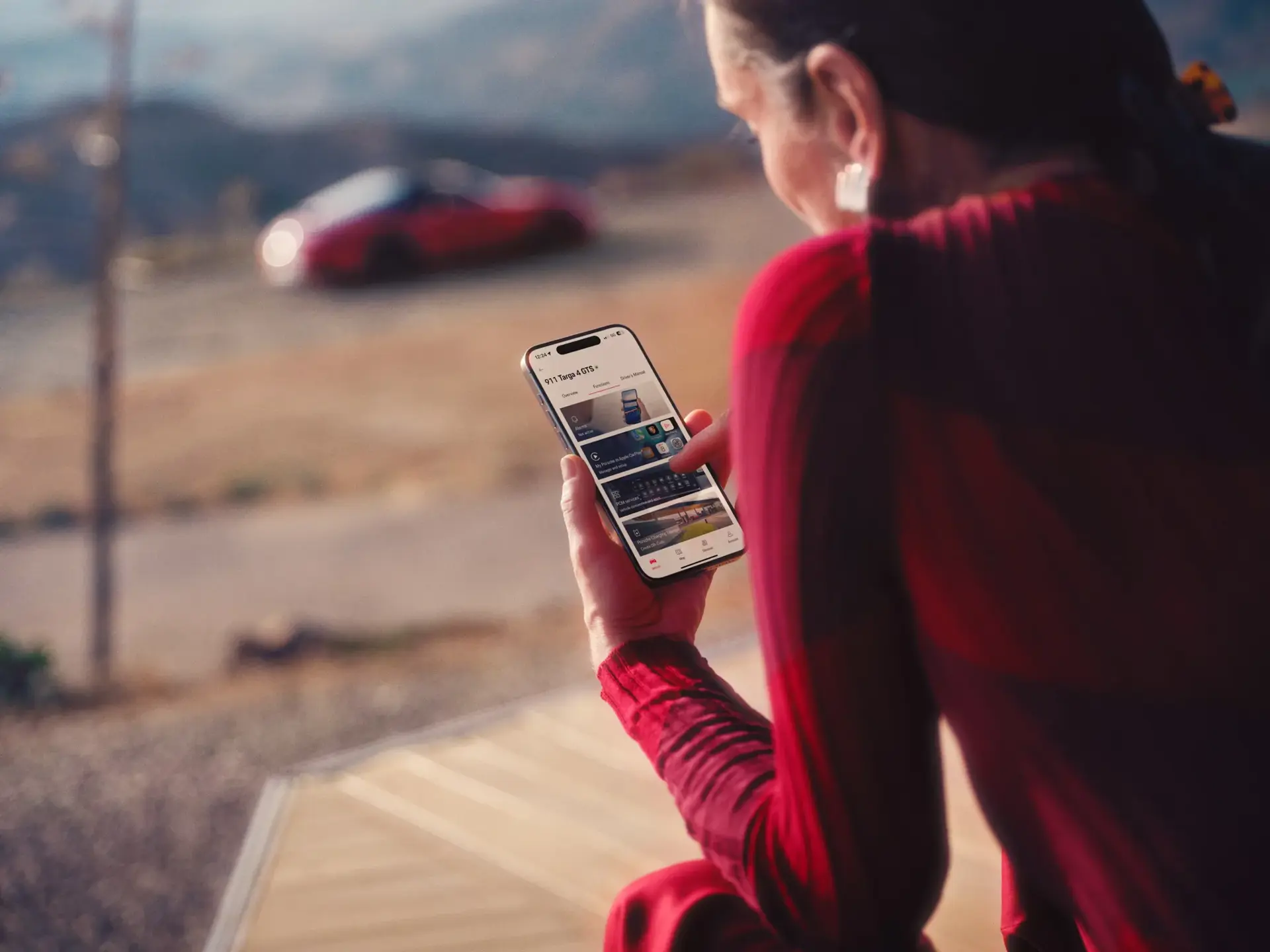 En person i rødt tøj sidder udendørs og bruger en smartphone-app til en Porsche, mens en rød sportsvogn ses sløret i baggrunden.