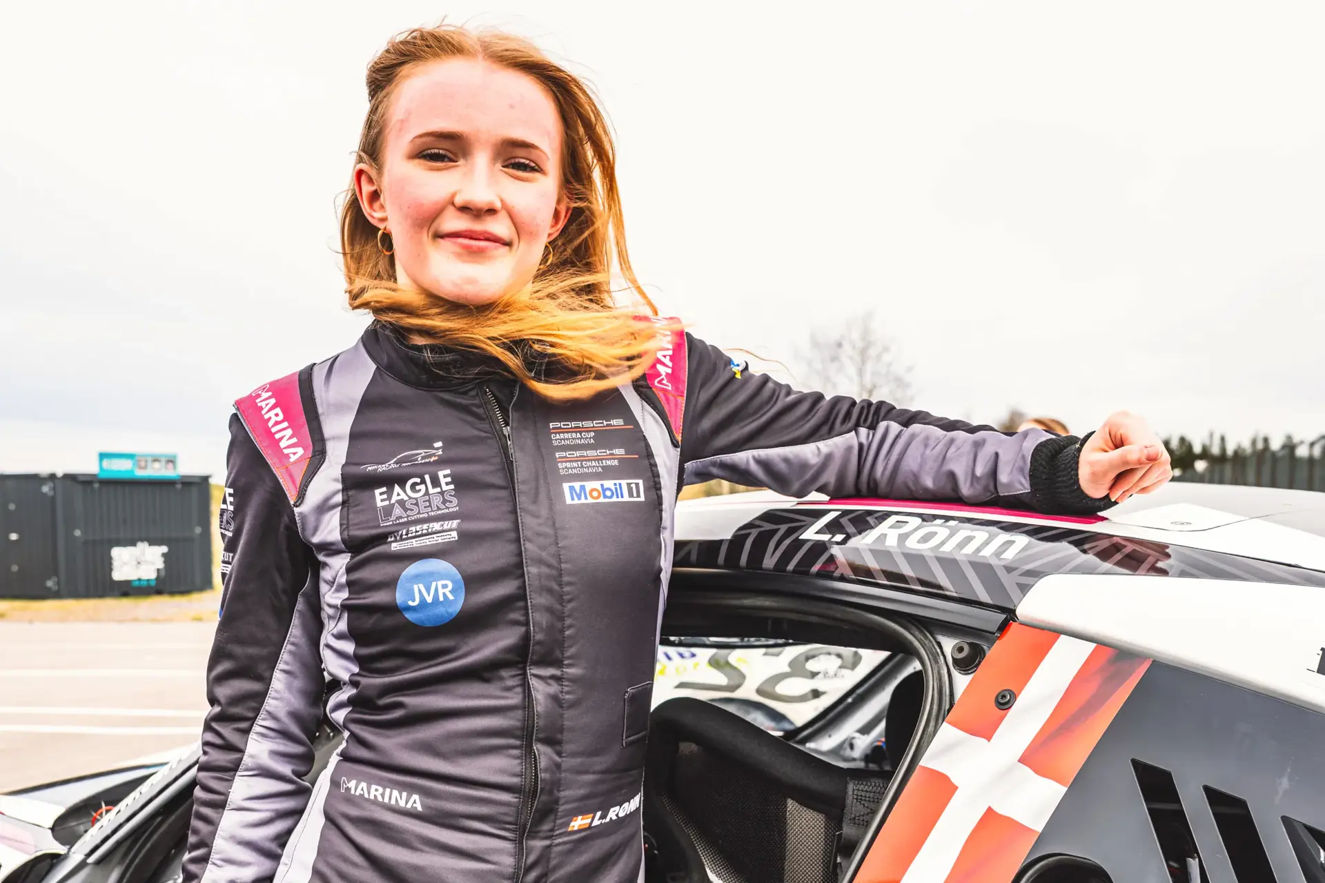 Racerkører i køredragt står ved siden af sin bil på banen med hånden hvilende på taget.