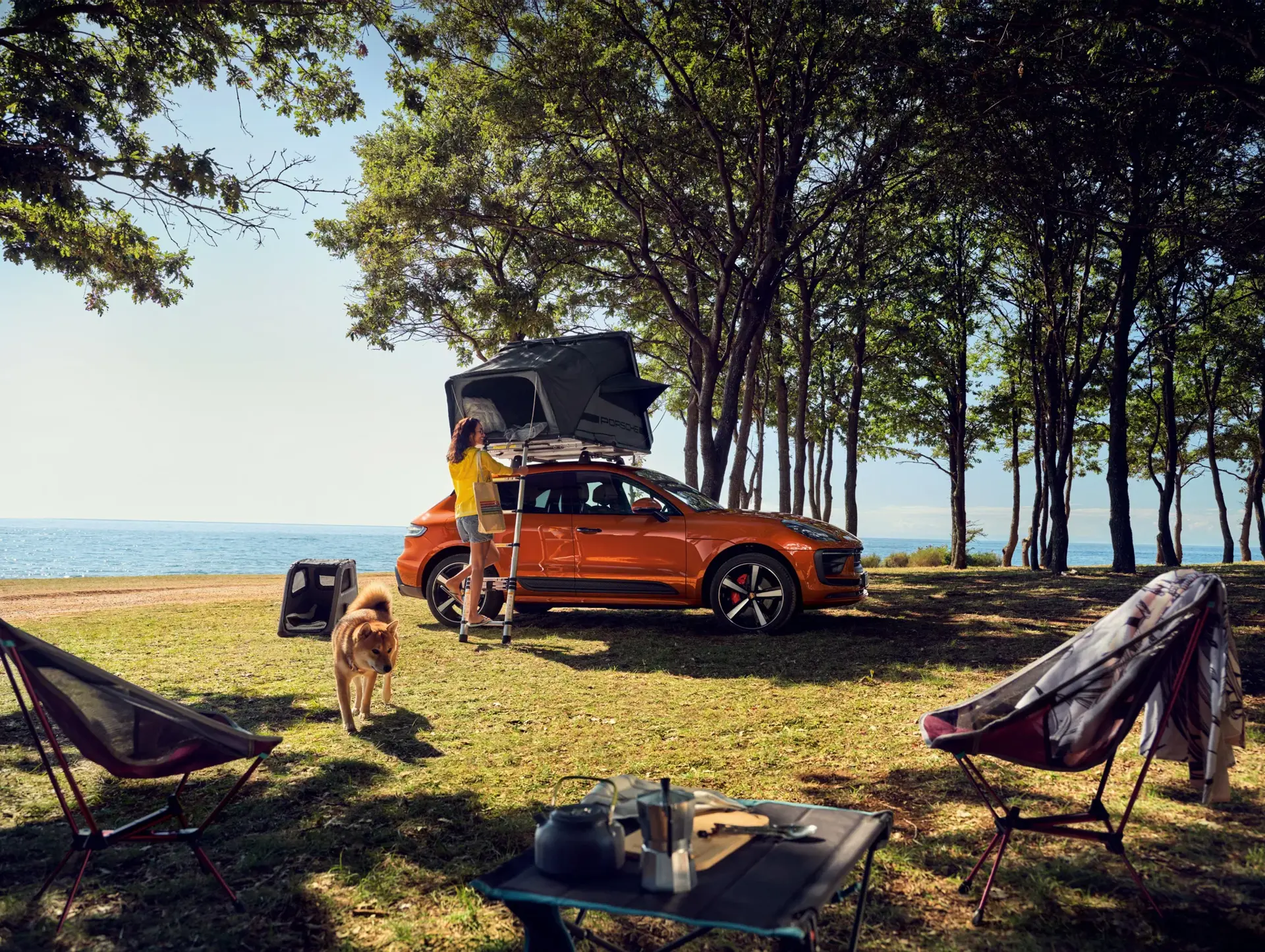 En kvinde camperer ved stranden med et orange SUV med tagtelt, omgivet af træer, campingstole og sin hund.