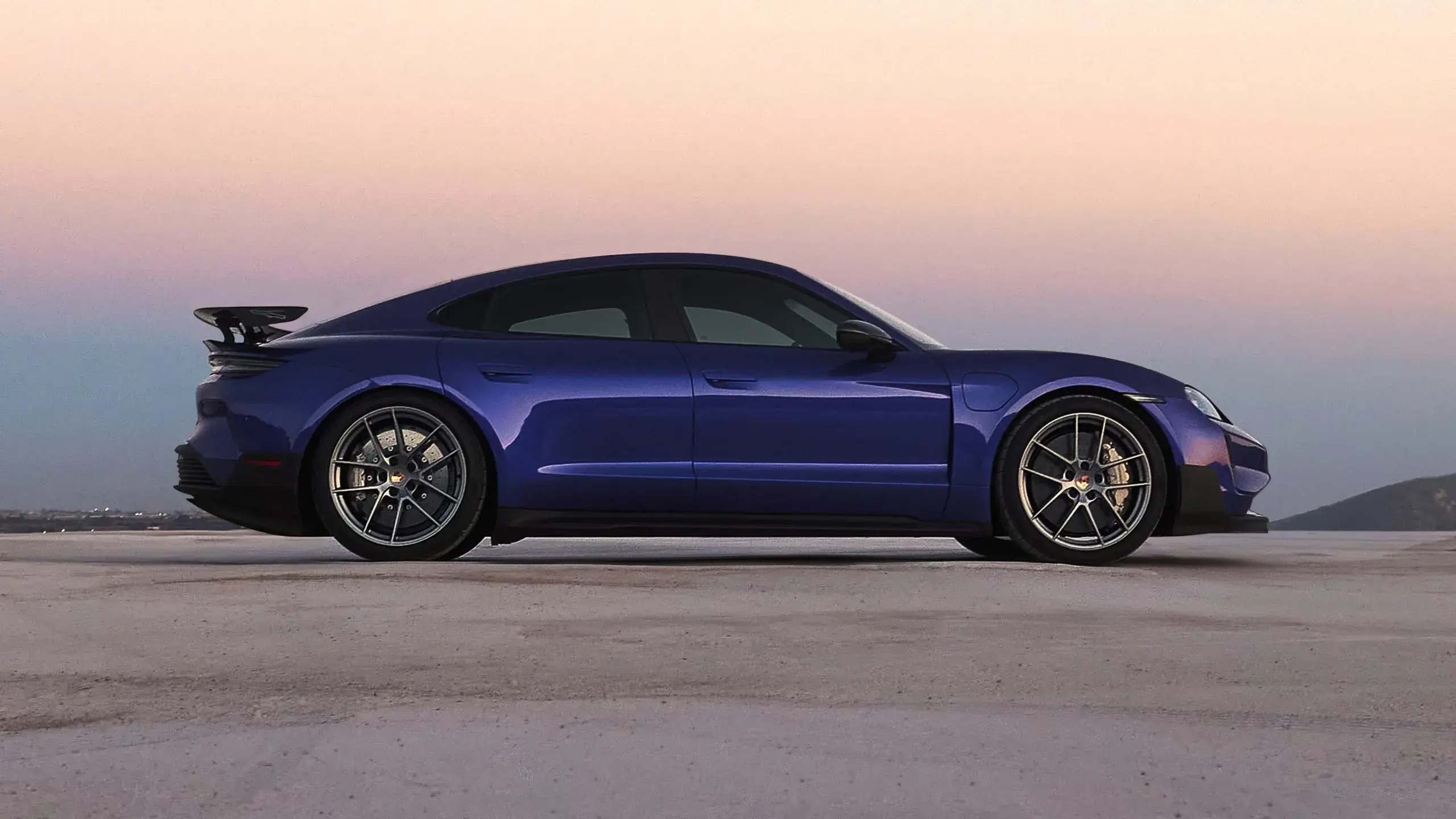 Bagfra-vinkel af en blå Porsche Taycan Turbo GT med stor hækspoiler i fart på en solrig vej.
