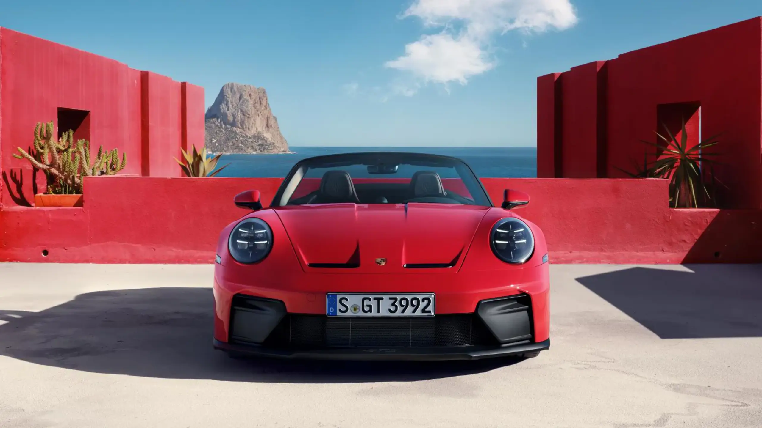 Rød Porsche 911 cabriolet parkeret ved moderne røde betonmure med udsigt over havet.