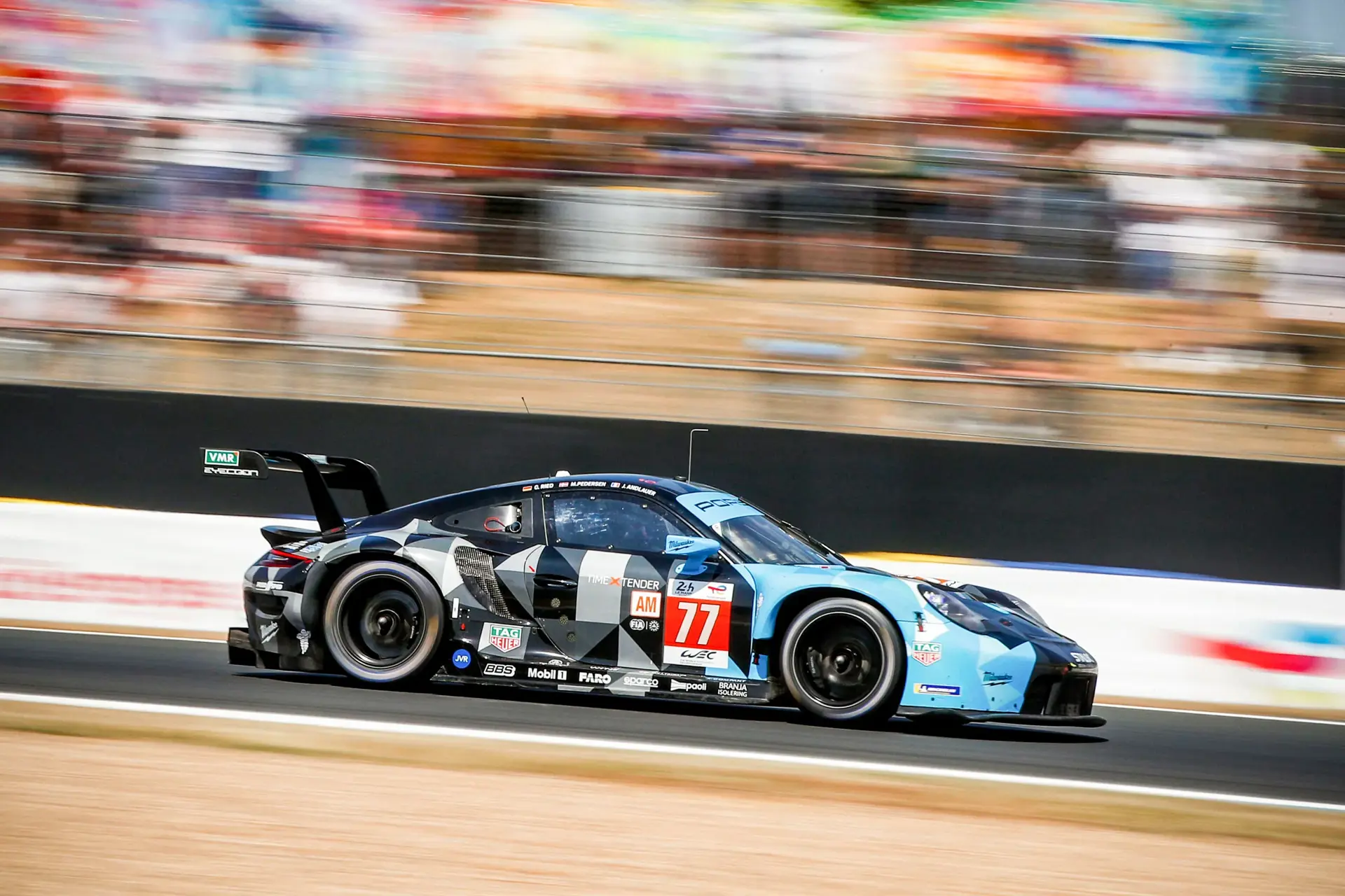 En blå og sort Porsche-racerbil med nummer 77 kører i høj fart på en racerbane med sløret baggrund.